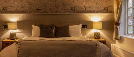 A bedroom with a patterned wallpaper, a bed with a herringbone throw, two bedside lamps, and a window with curtains.