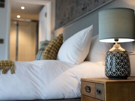 A neatly made bed with white linens and a patterned throw blanket, a bedside lamp, and a wooden nightstand with a drawer.