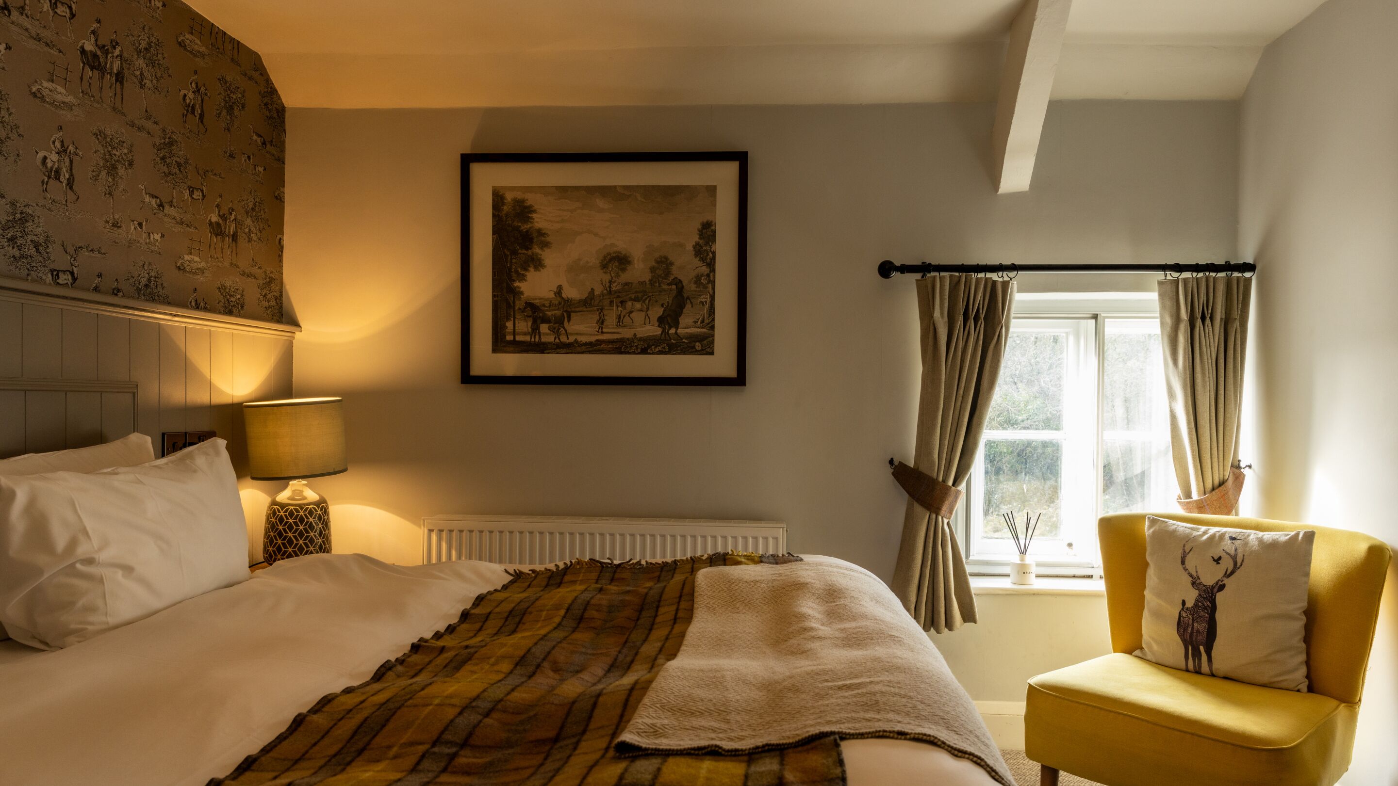 A bedroom with a bed, a bedside lamp, a framed picture on the wall, a window with curtains, and a chair with a cushion.