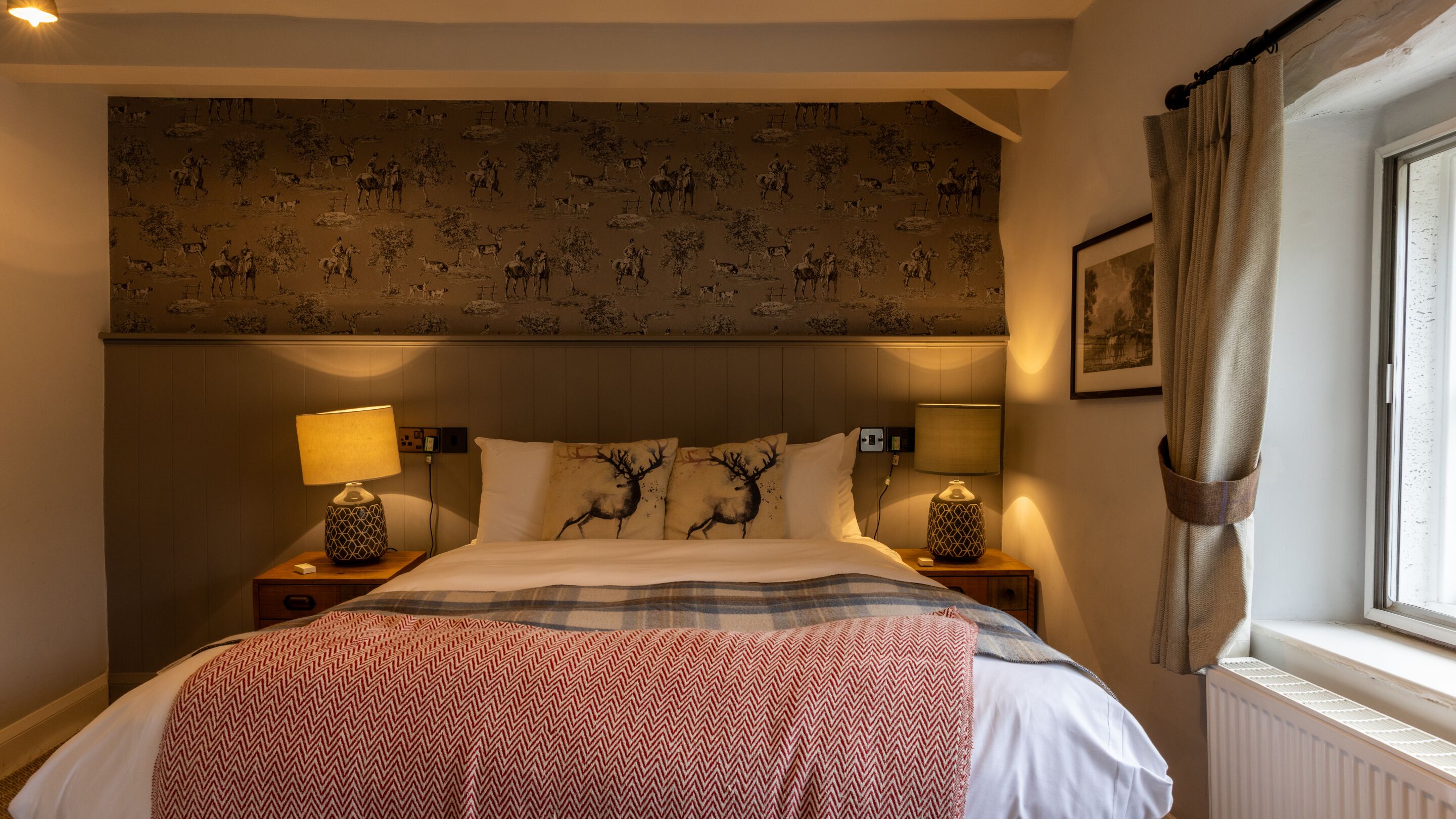 A hotel room with a bed, two bedside lamps, a headboard with a patterned wallpaper, and a window with curtains.