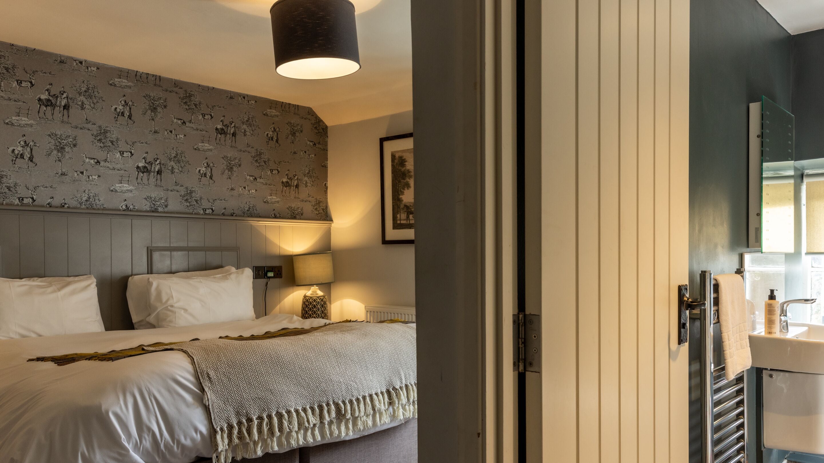 Habitación de hotel con cama, lámpara de pared, mesita de noche y baño visible a través de una puerta abierta.