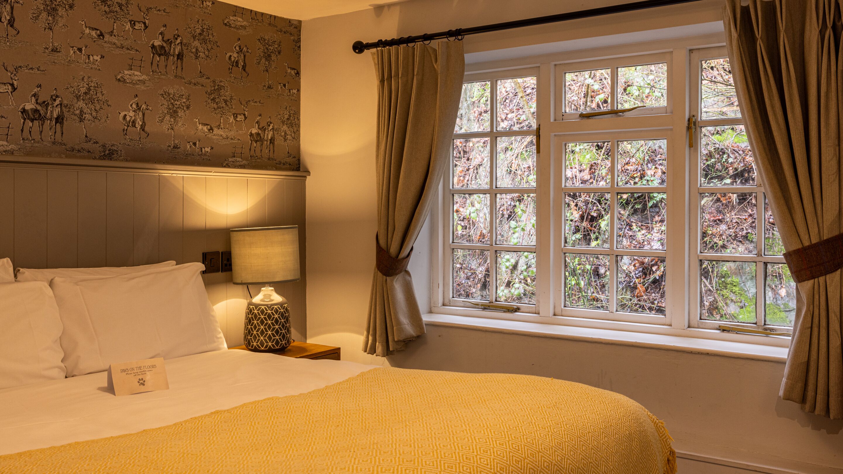 Una habitación de hotel con cama, mesita de noche, lámpara, ventana con cortinas y papel tapiz con motivos naturales.