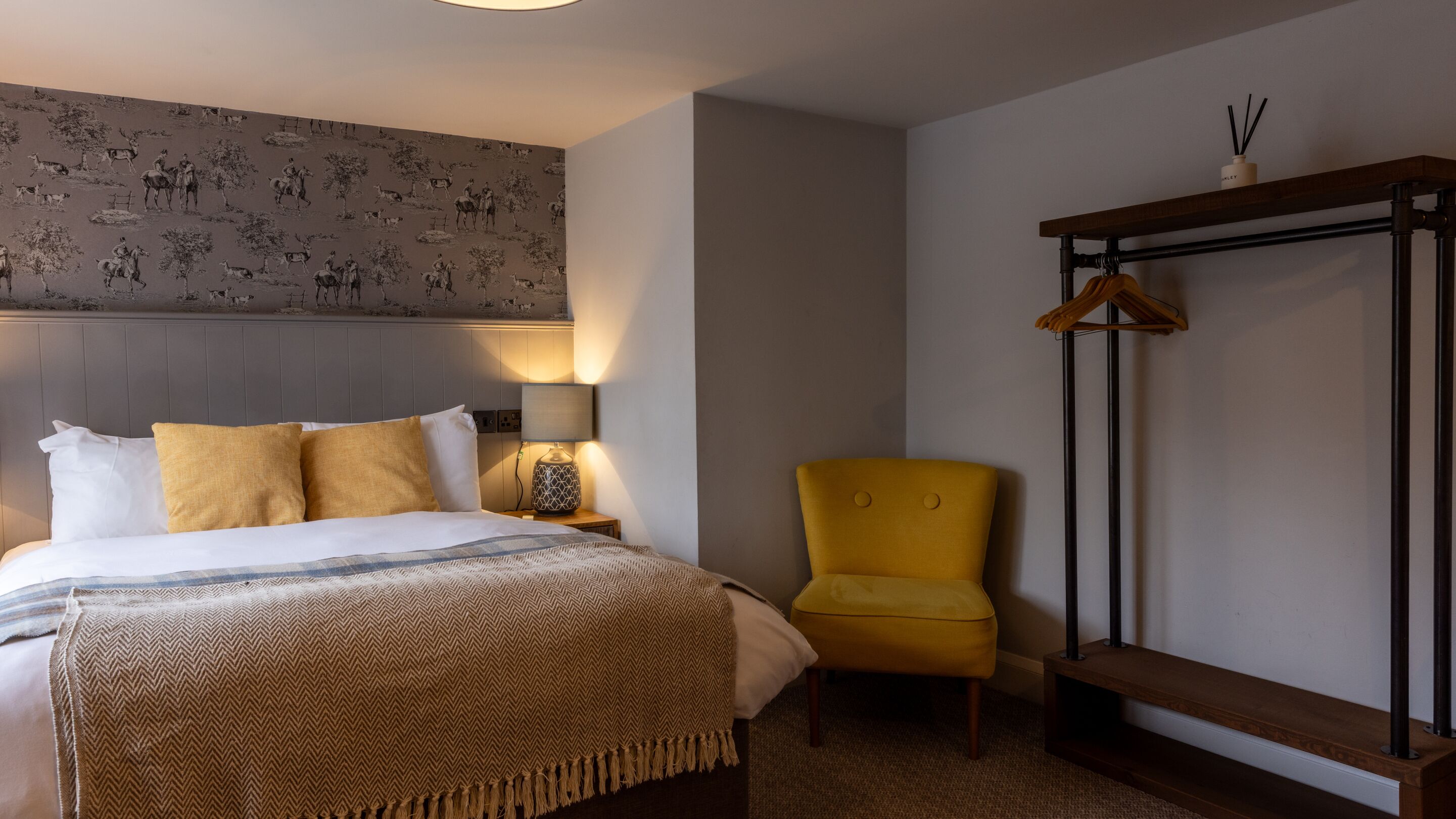 Habitación de hotel con cama, una silla, perchero y un papel tapiz con diseño.