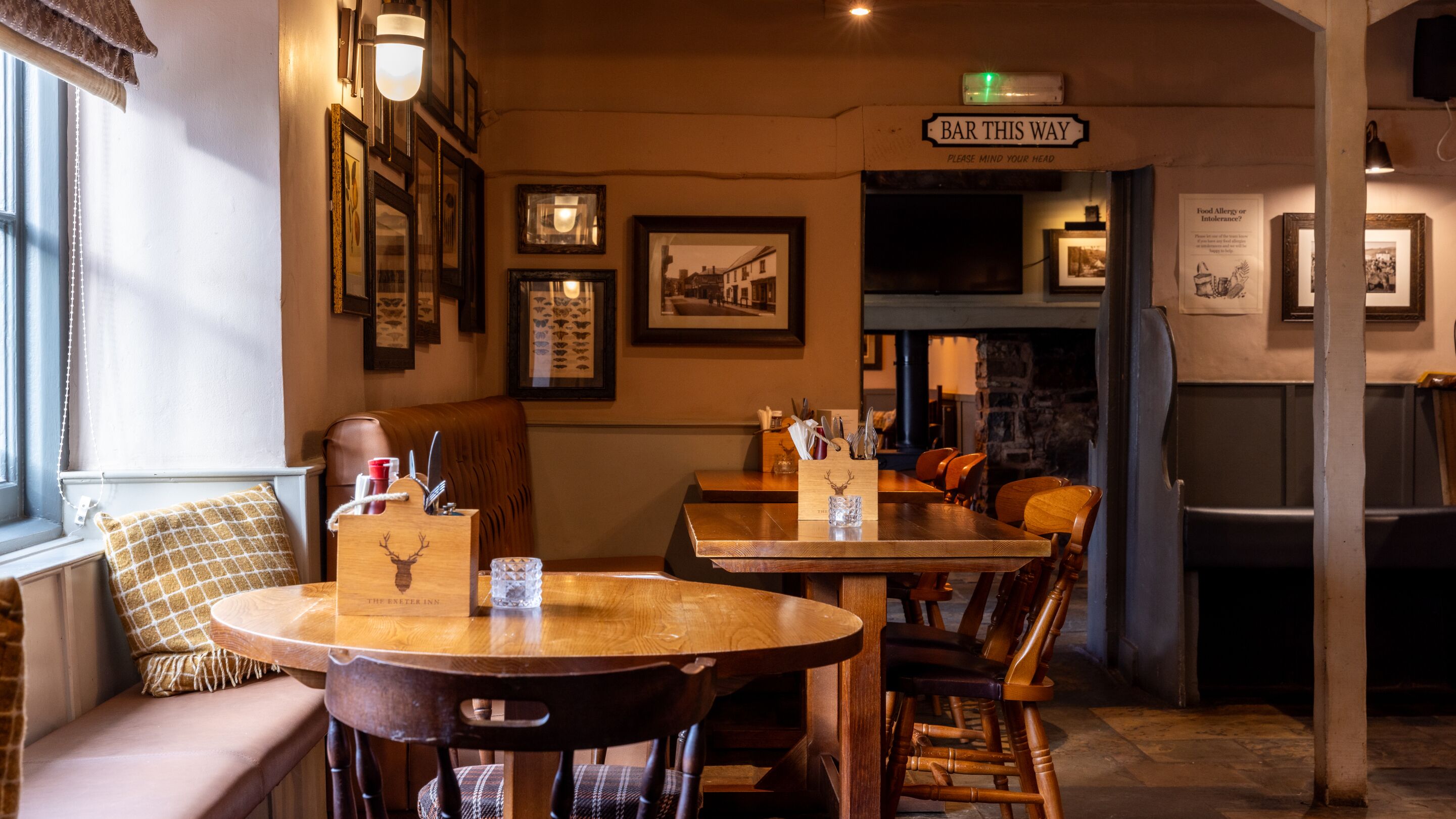 A cozy bar area with wooden tables and chairs, framed pictures on the walls, and a sign reading 'BAR THIS WAY'.