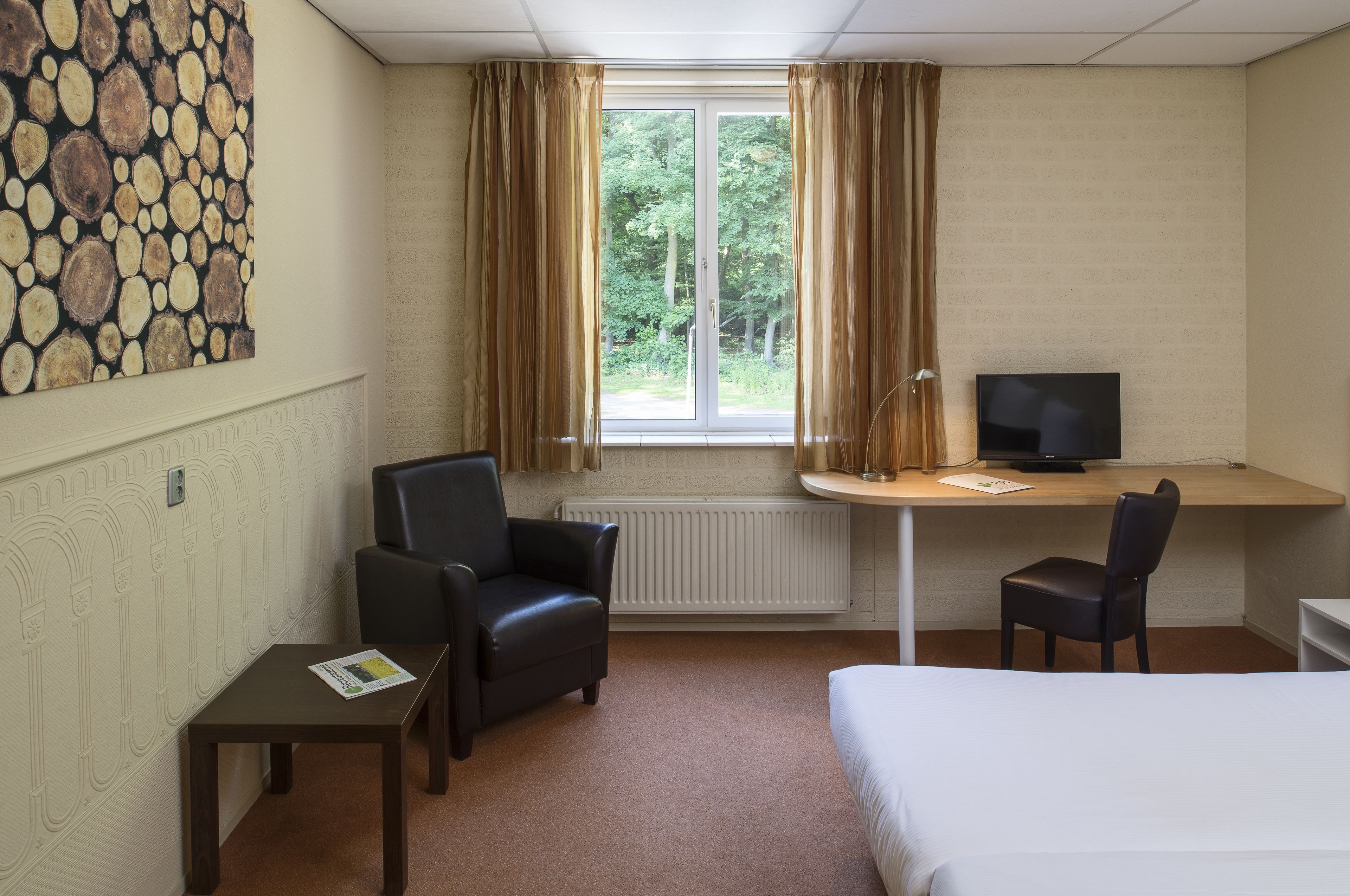 A hotel room with a bed, a desk with a computer, two chairs, and a window with curtains.