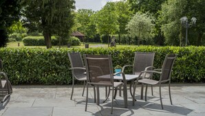 Un patio al aire libre con cuatro sillas de mimbre y una mesa, rodeado de un seto y árboles.