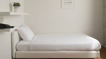 A small, white, single-bed room with a brown carpet, a white wall, a shelf with a plant, and a framed picture.