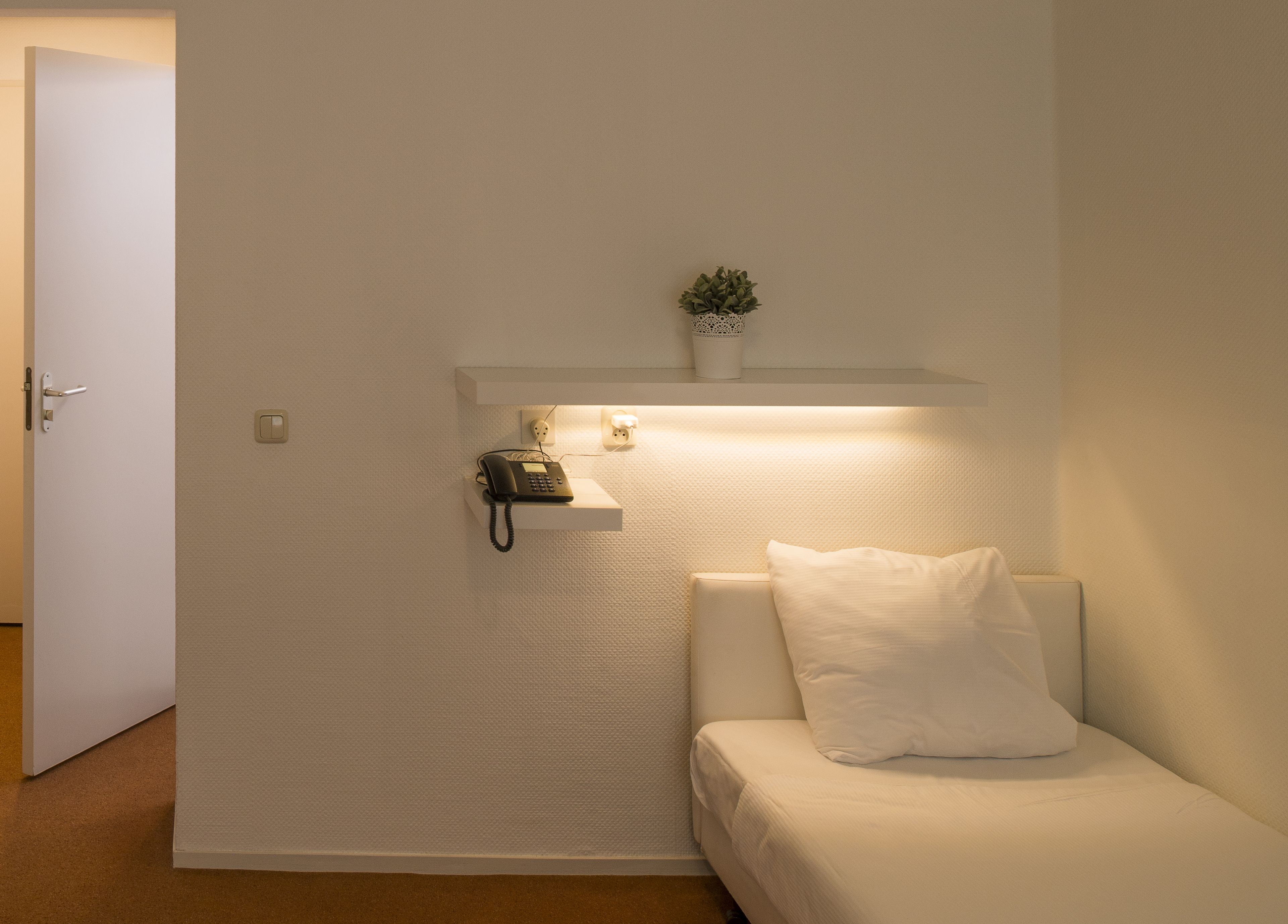 Una stanza dal design minimalista con una parete bianca, un piccolo ripiano con un telefono e una pianta, e un divano bianco con un cuscino.