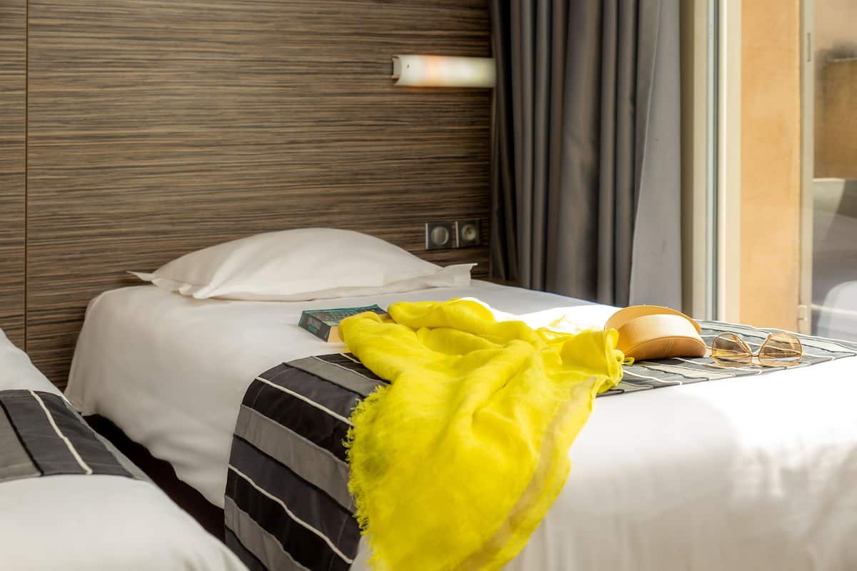 A hotel room with a wooden headboard, a bed with white bedding, a striped pillow, a yellow throw blanket, and a pair of glasses on the bed.