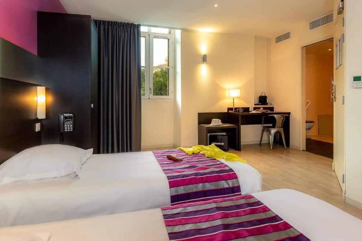 A hotel room with two beds, a desk, a chair, and a window.