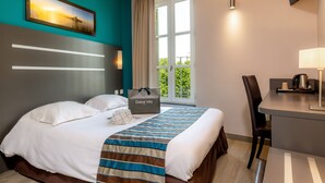 Una habitación de hotel con cama, escritorio, silla, ventana con cortinas, un cuadro en la pared y un bolso Dolce Vita sobre la cama.