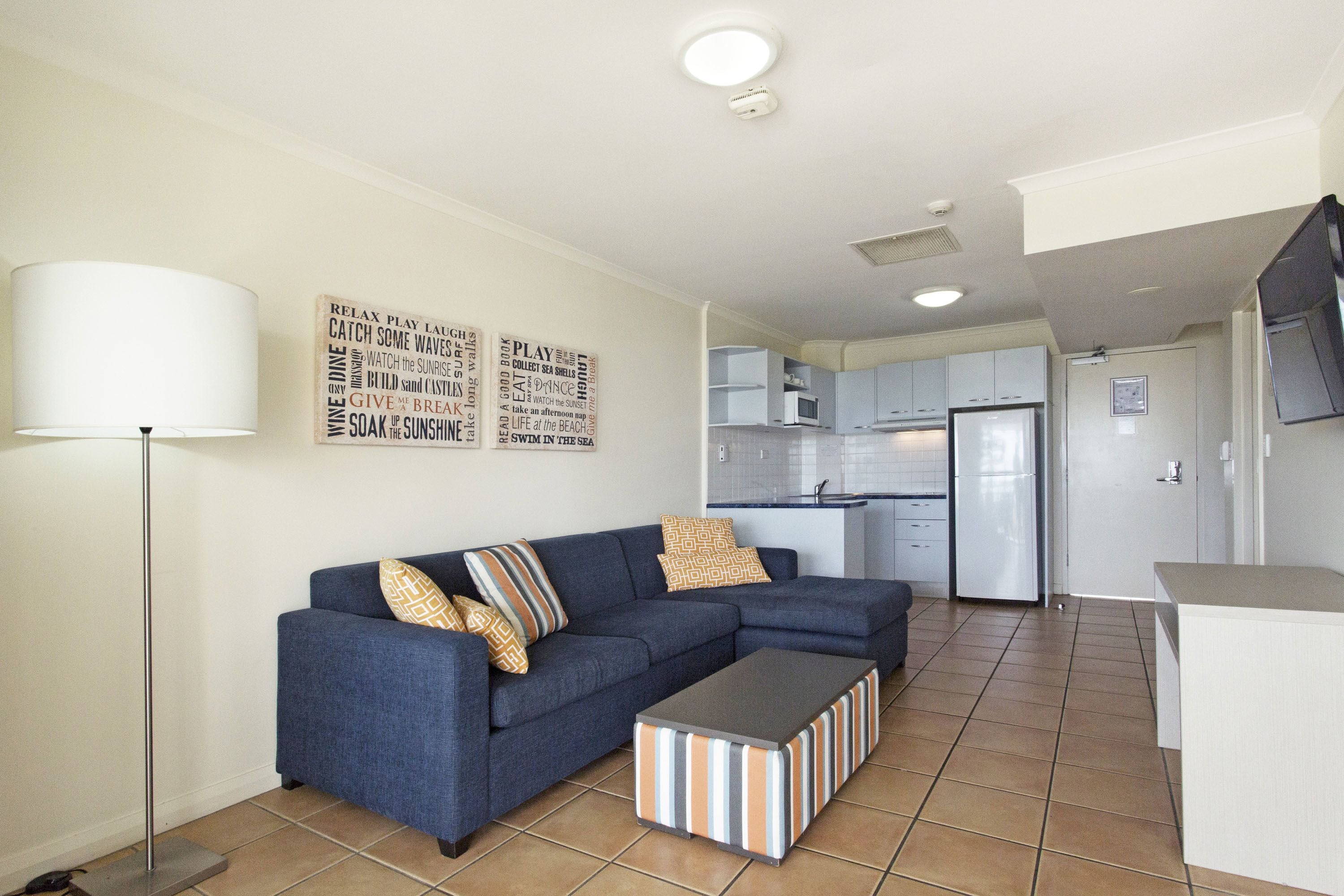 A modern living room with a blue sofa, striped ottoman, and a lamp. A kitchen is visible in the background with white cabinets and a refrigerator.