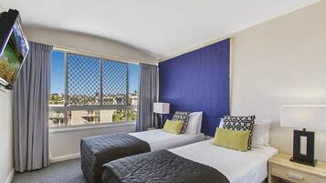 Une chambre d’hôtel avec deux lits, un téléviseur fixé au mur, une fenêtre donnant sur des immeubles et un mur d’accent bleu.