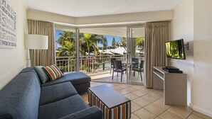 A modern living room with a sofa, a coffee table, and a TV. It has a balcony with a view of palm trees and a pool.