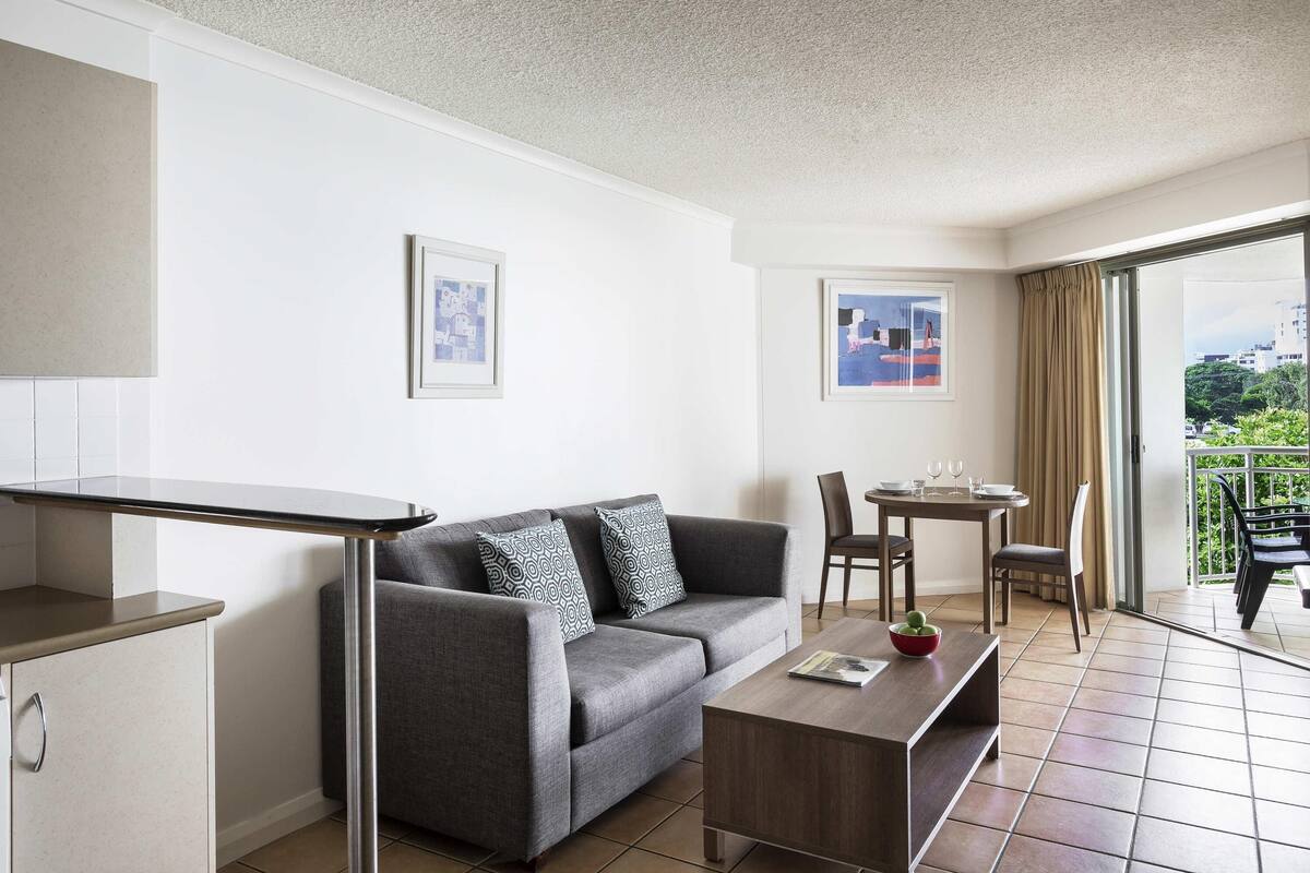 A modern living room with a grey sofa, a wooden coffee table, a dining table with chairs, and a balcony with a view.