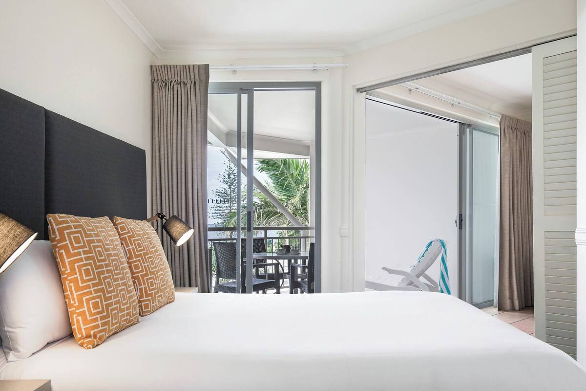 A hotel room with a large bed, a balcony with a table and chairs, and a view of greenery.