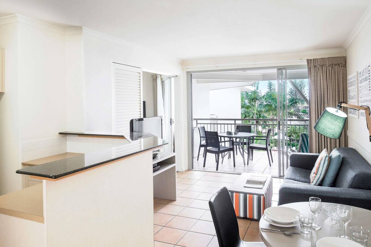A modern kitchen with a dining area, a balcony with a view of palm trees, and a grey sofa with a striped cushion.