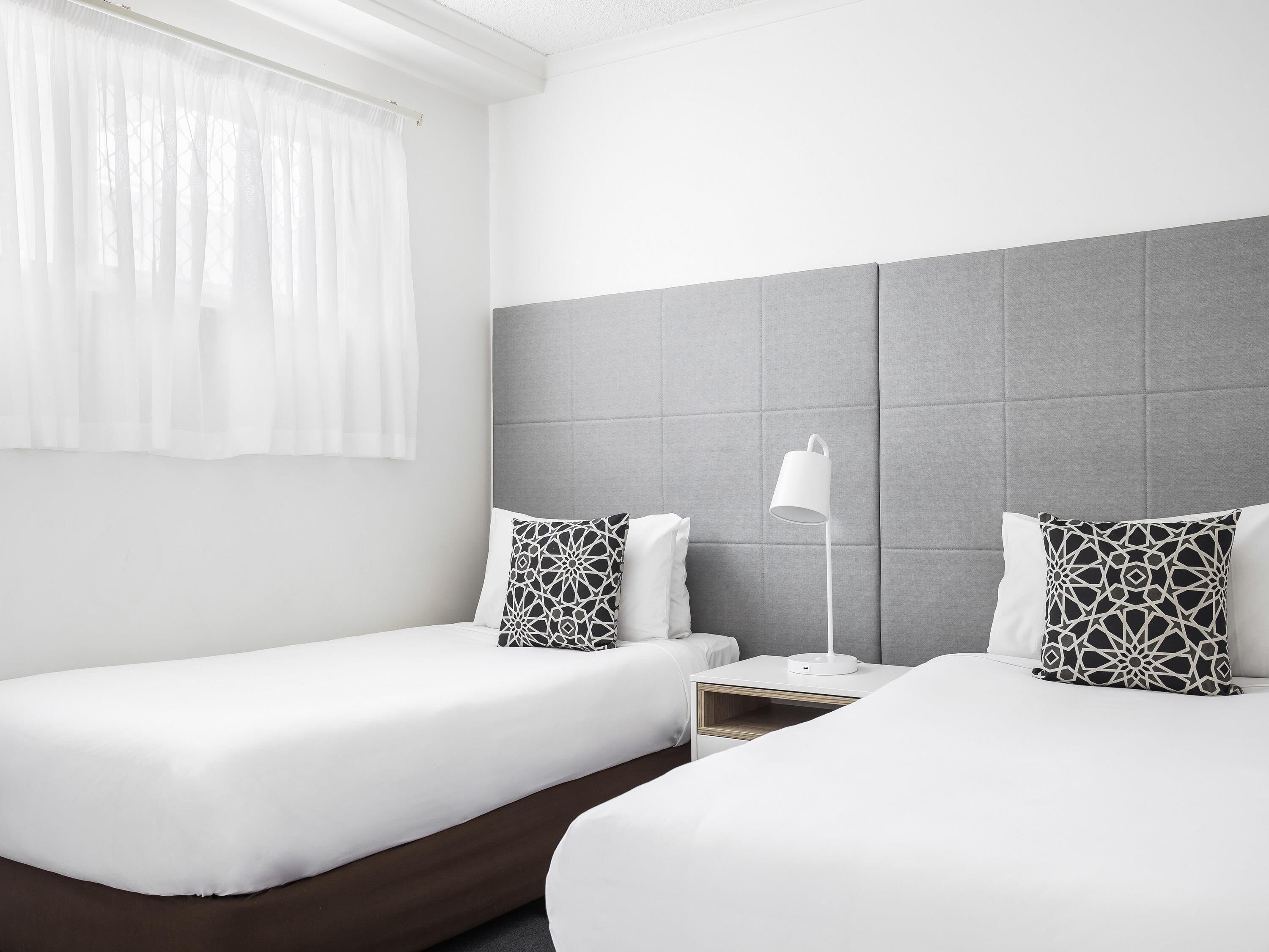 Une chambre d’hôtel avec deux lits simples, une table de chevet avec une lampe et une fenêtre avec des rideaux légers.