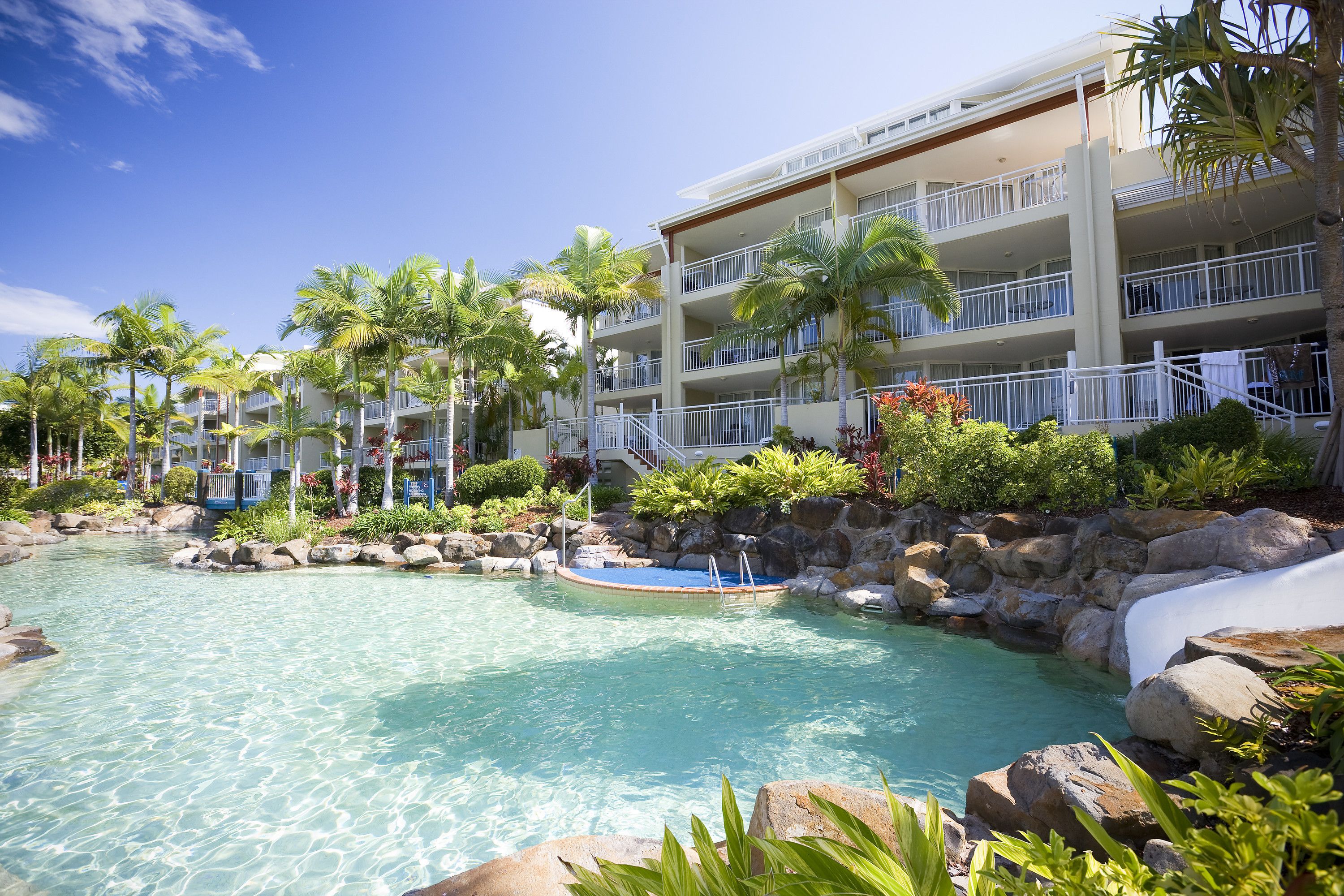 A resort with a clear pool, palm trees, and a multi-story building with balconies.