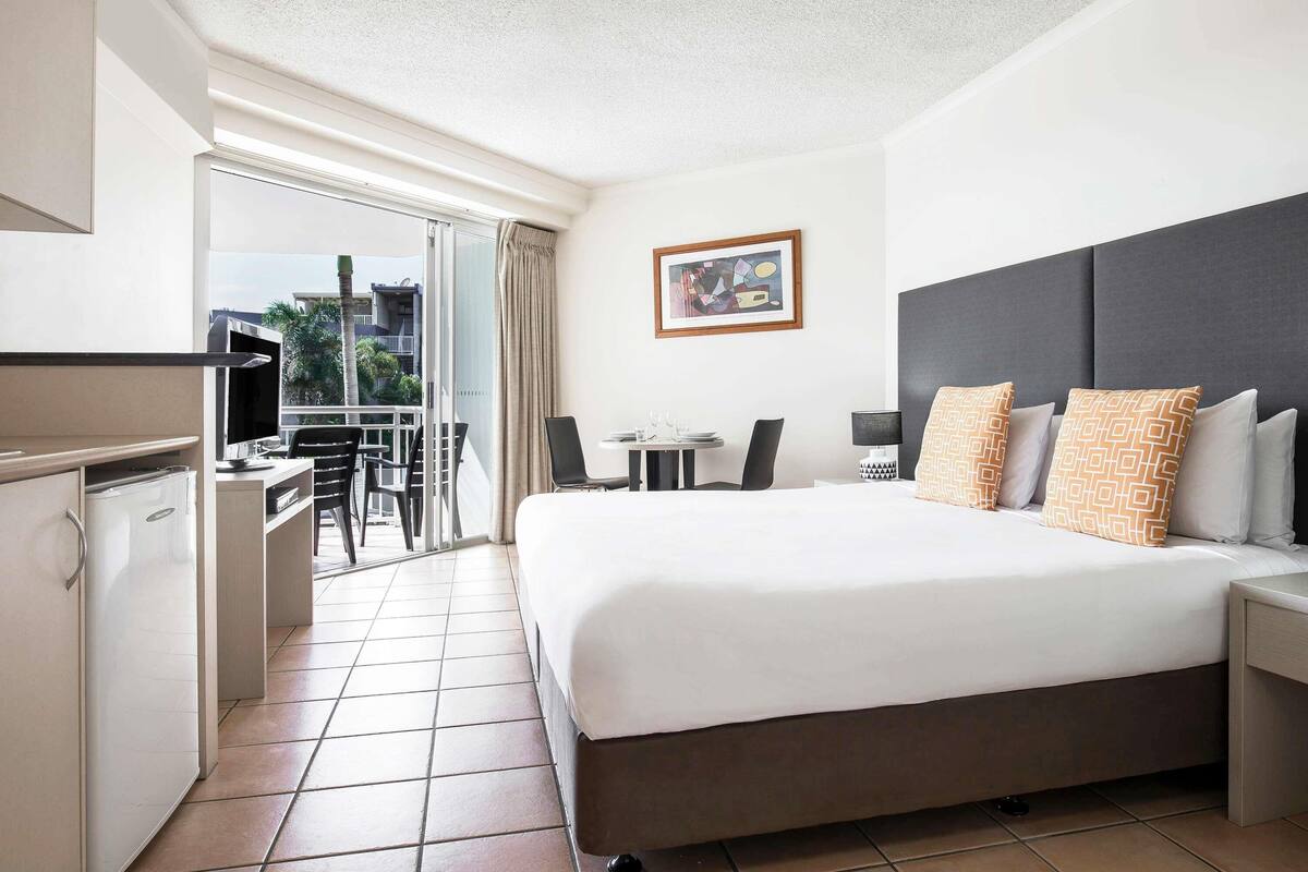 A hotel room with a bed, a desk, a chair, a small table, a TV, and a balcony with a view.