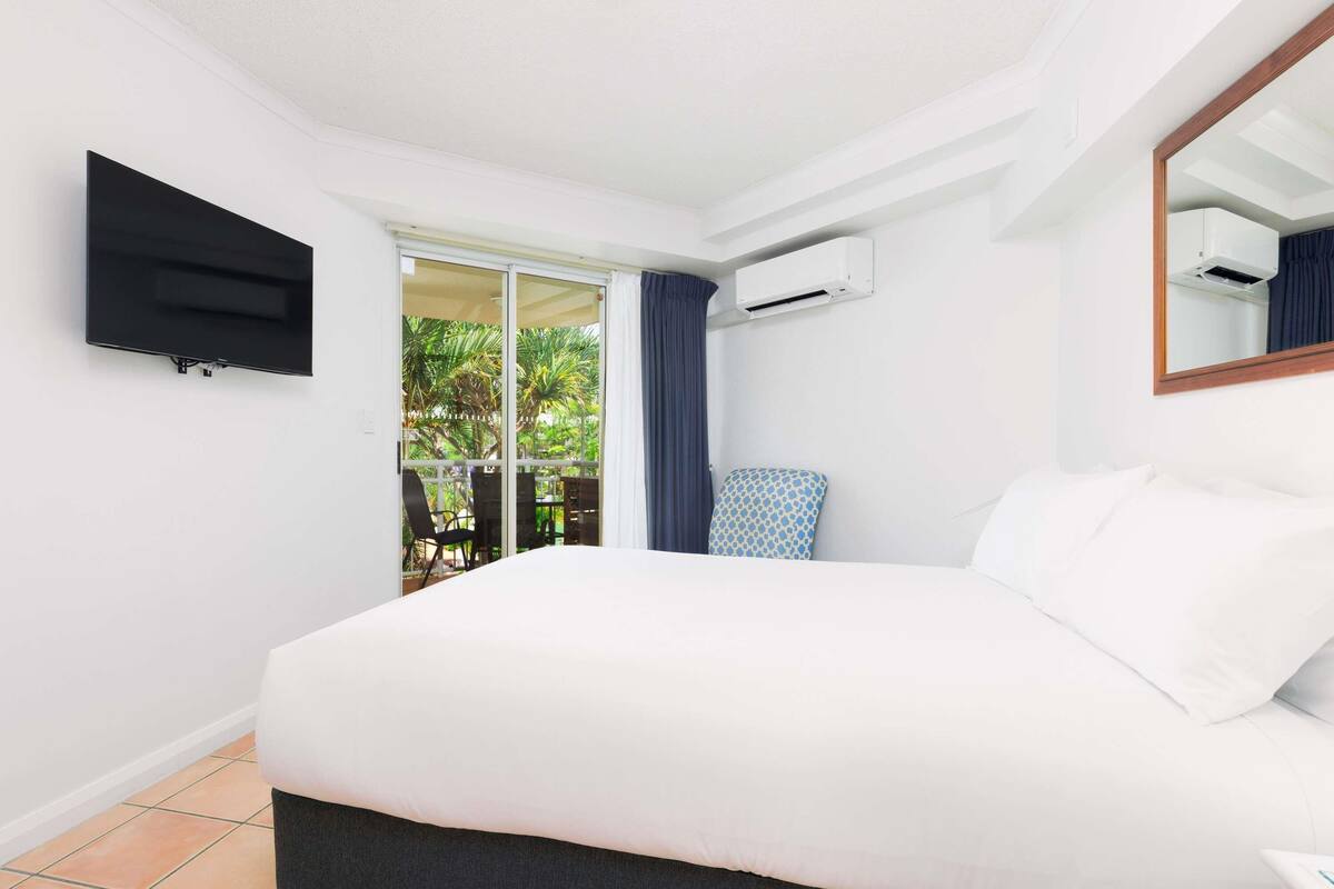 A hotel room with a bed, a TV mounted on the wall, an air conditioning unit, and a view of the outdoors.
