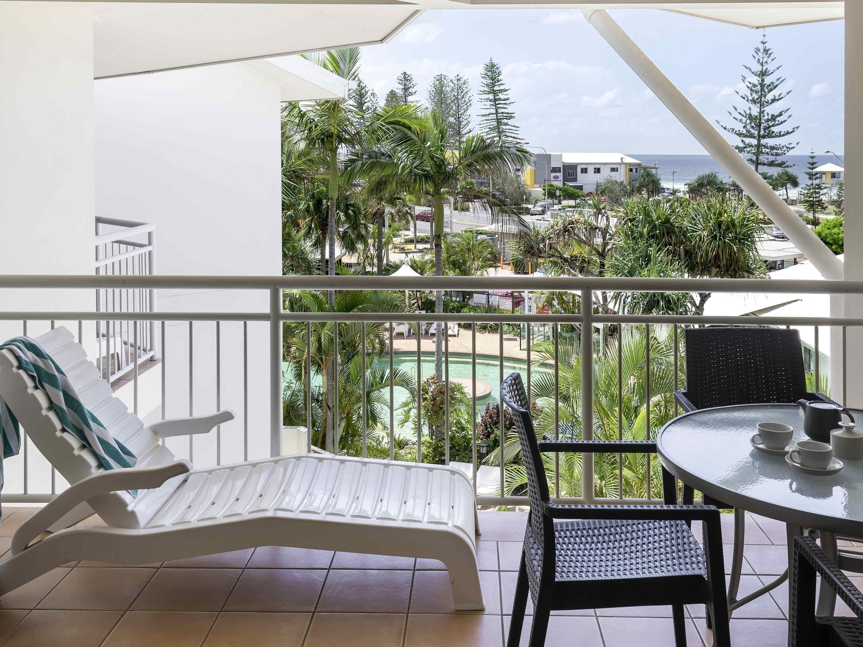 A balcony with a white lounge chair, a round table with chairs, and a view of a pool and buildings.