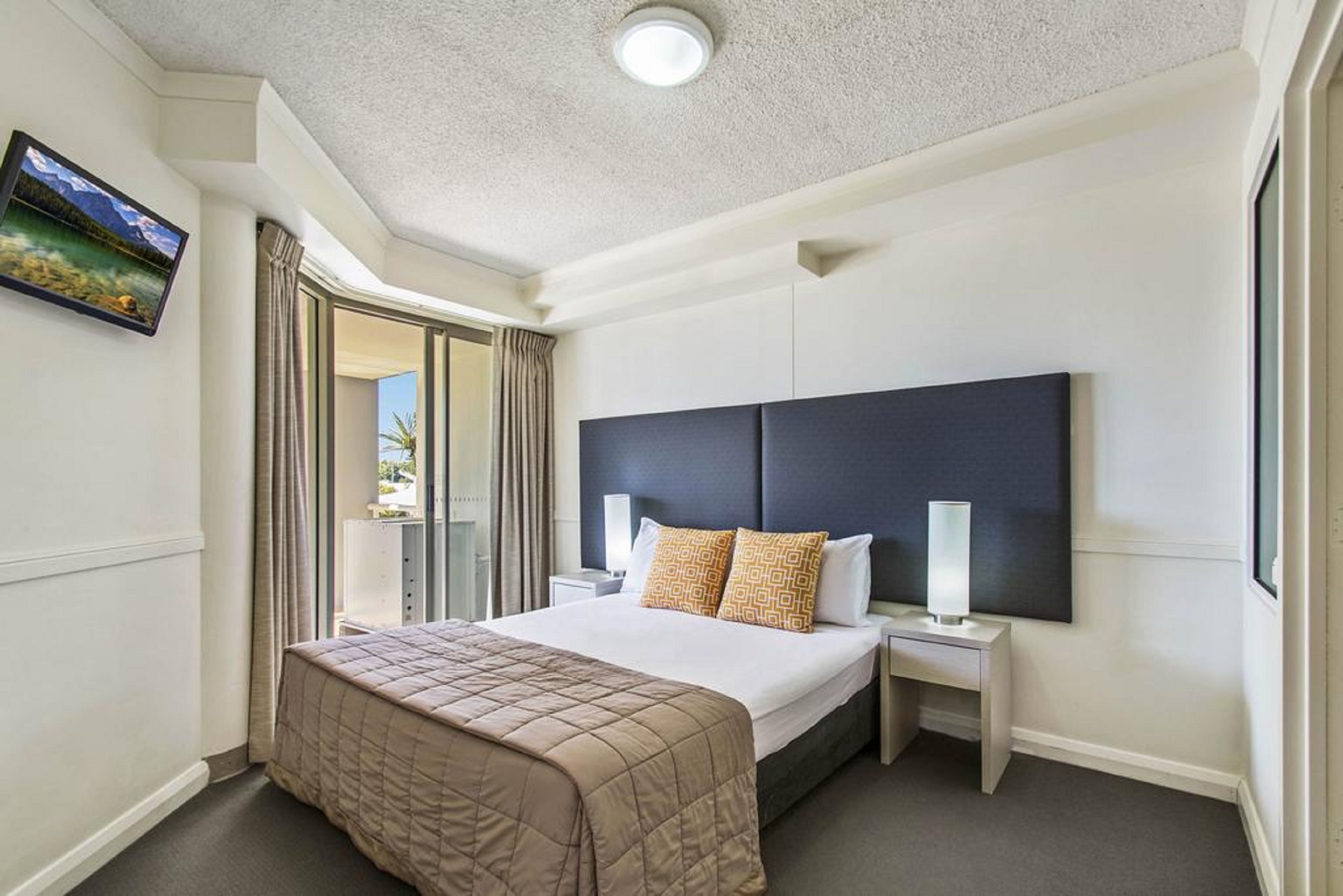 Quarto de hotel com uma cama grande, cabeceira, mesa de cabeceira, televisão de tela plana na parede e uma janela com cortinas.