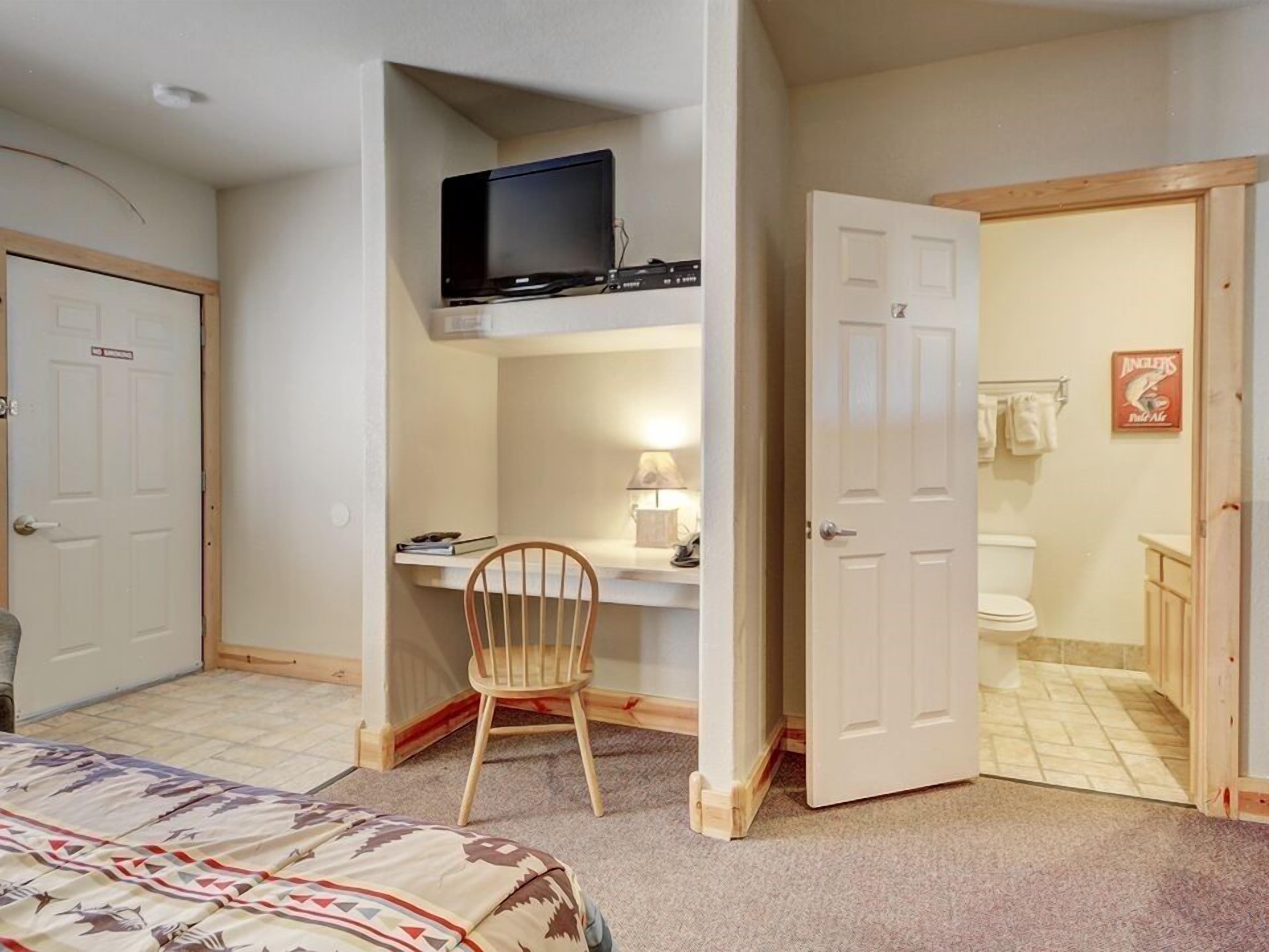 A bedroom with a bed, a desk with a chair, a television, and a bathroom visible through an open door.
