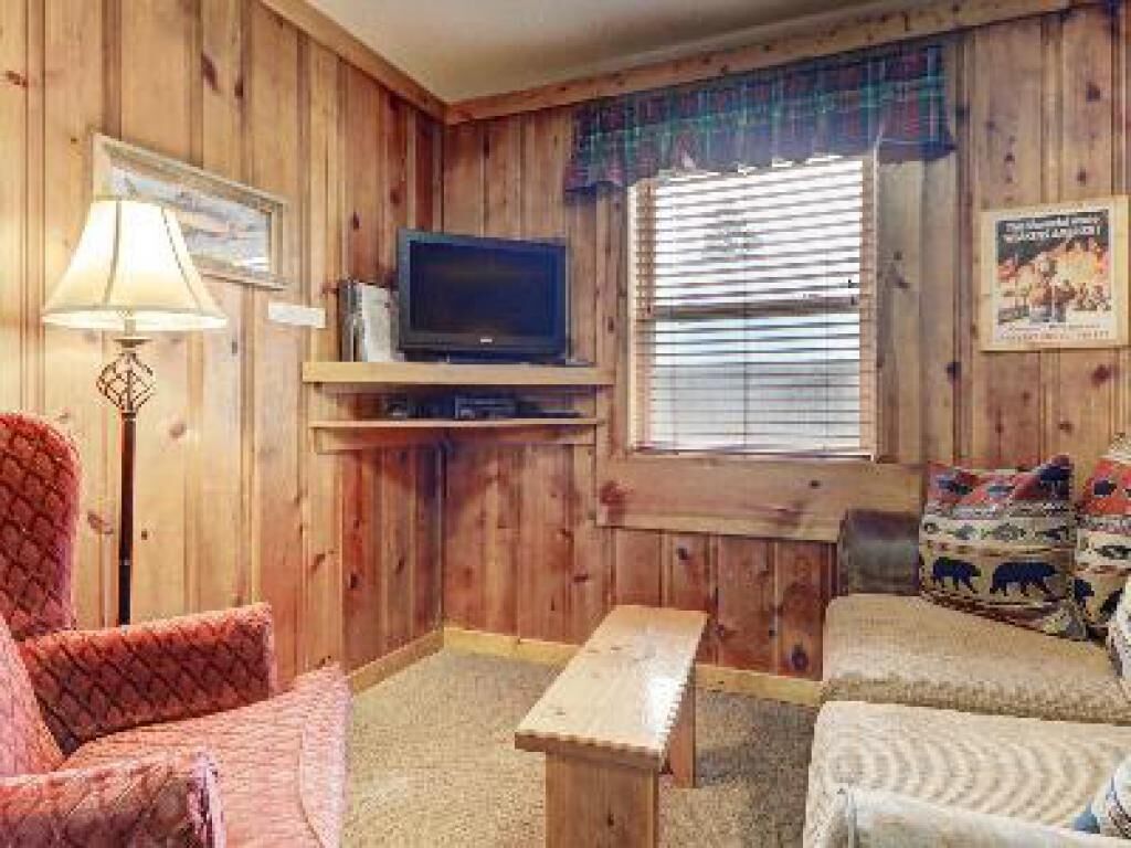 A cozy cabin interior with wooden walls, a television on a shelf, a bench, and a couch with patterned cushions.