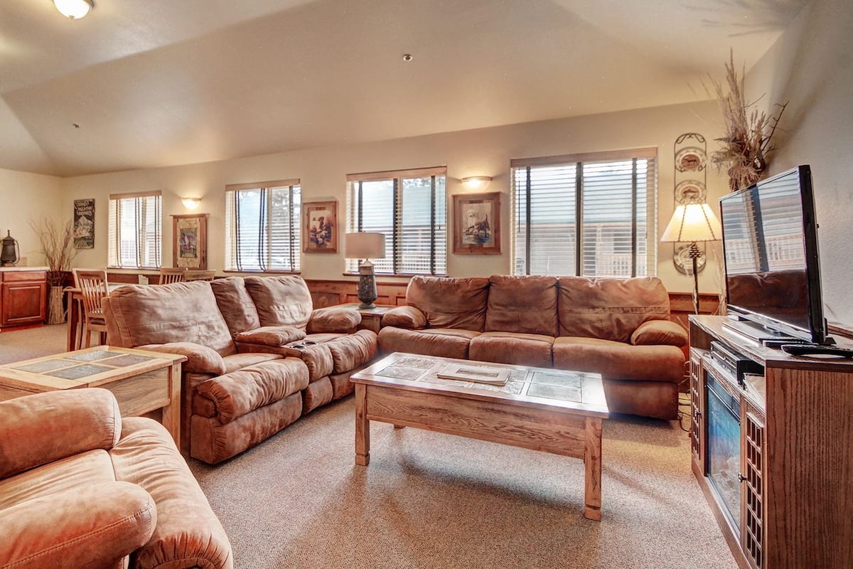 A spacious living room with a large brown leather sofa, a wooden coffee table, and a television on a wooden cabinet. There are multiple windows with blinds, a kitchen area in the background, and a dining table with chairs.