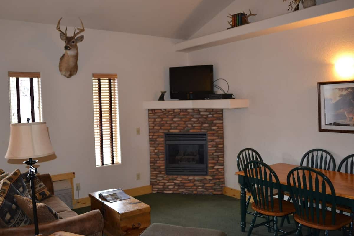 A living room with a stone fireplace, a mounted deer head, a television on a mantel, and a dining area with wooden chairs and a table.