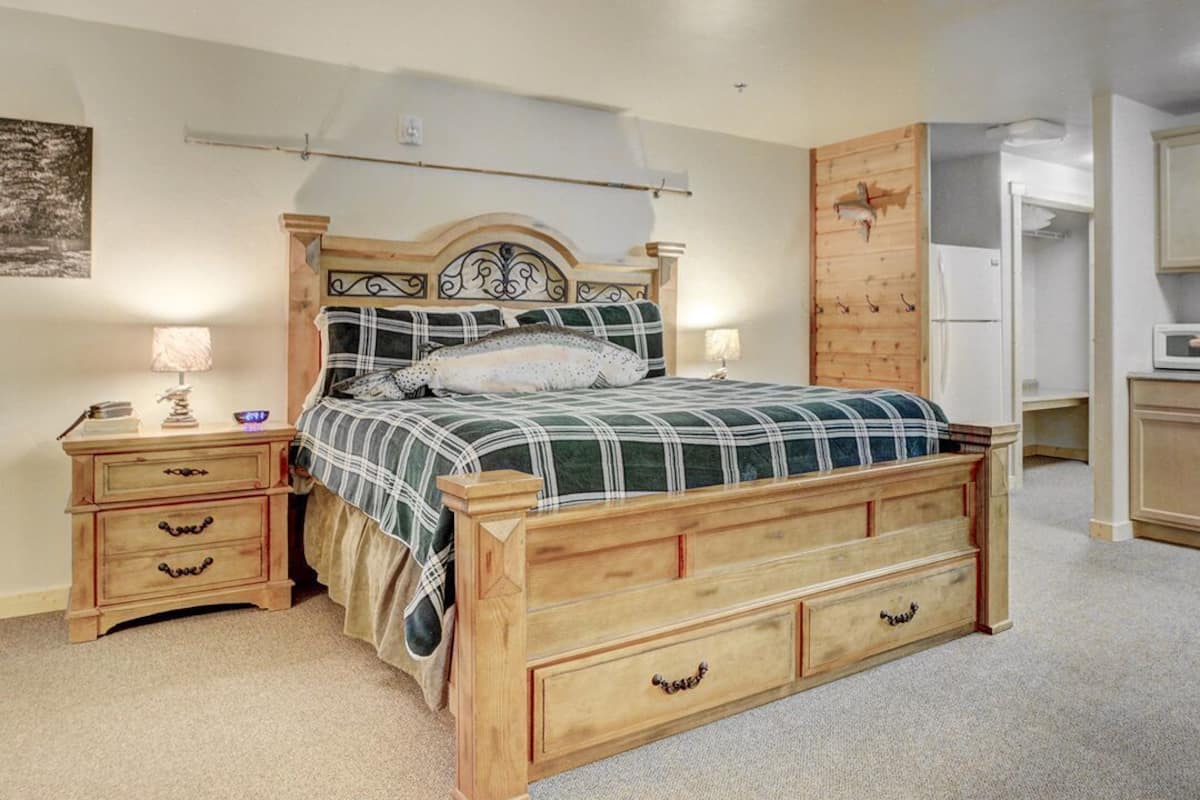 A bedroom with a wooden bed, bedside tables, a wooden wardrobe, and a kitchen area with cabinets and a microwave.