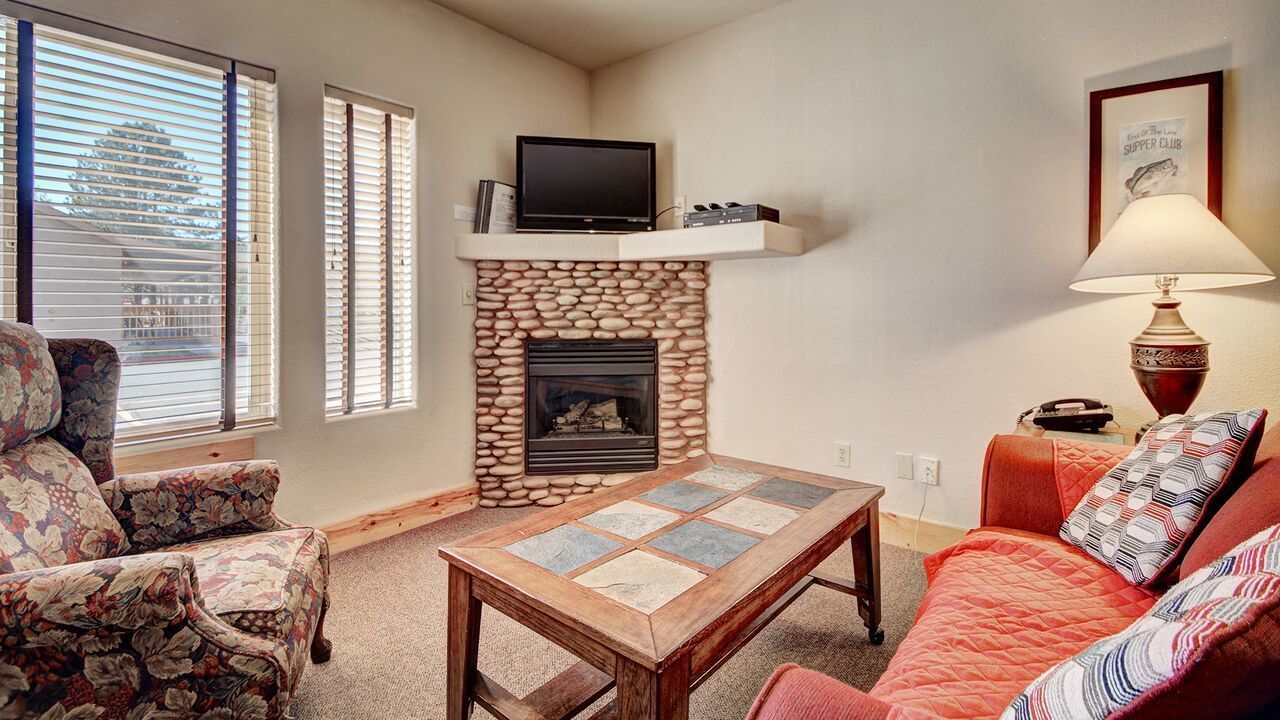 Una sala de estar con chimenea de piedra, un televisor en un estante encima, un sillón estampado de flores, un sofá rojo y una mesa de centro con motivos.