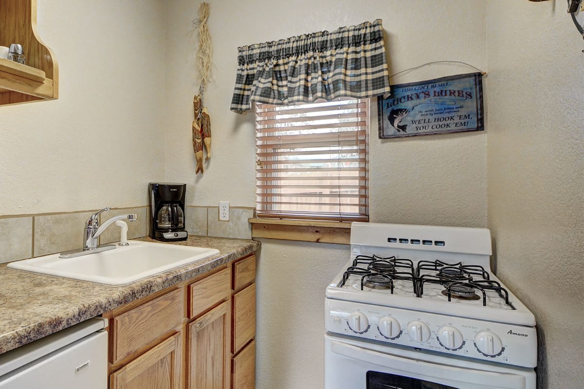 A compact kitchen with a white sink, a gas stove, wooden cabinets, and a window with blinds.