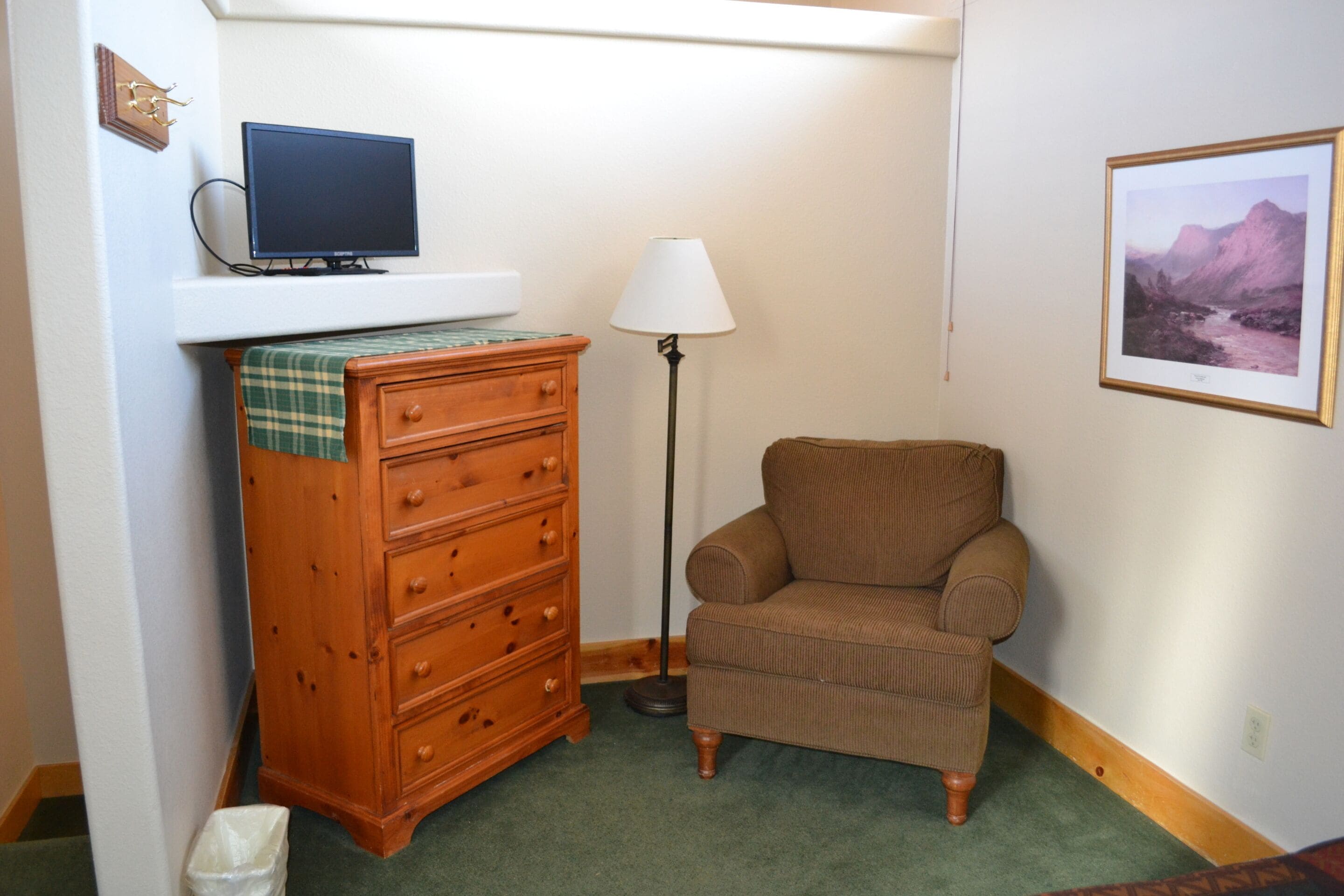 Una habitación con un aparador de madera, un sillón marrón, un televisor sobre una estantería, una lámpara de pie y un cuadro en la pared.