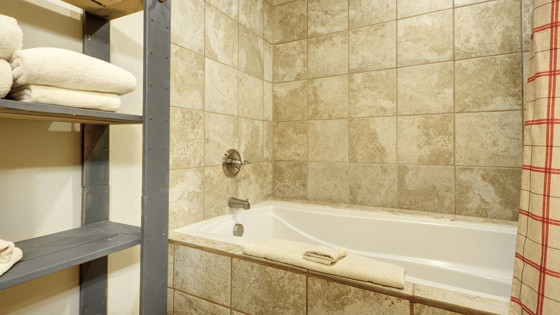 A bathroom with a bathtub, beige tiled walls, and a metal shelf with towels.
