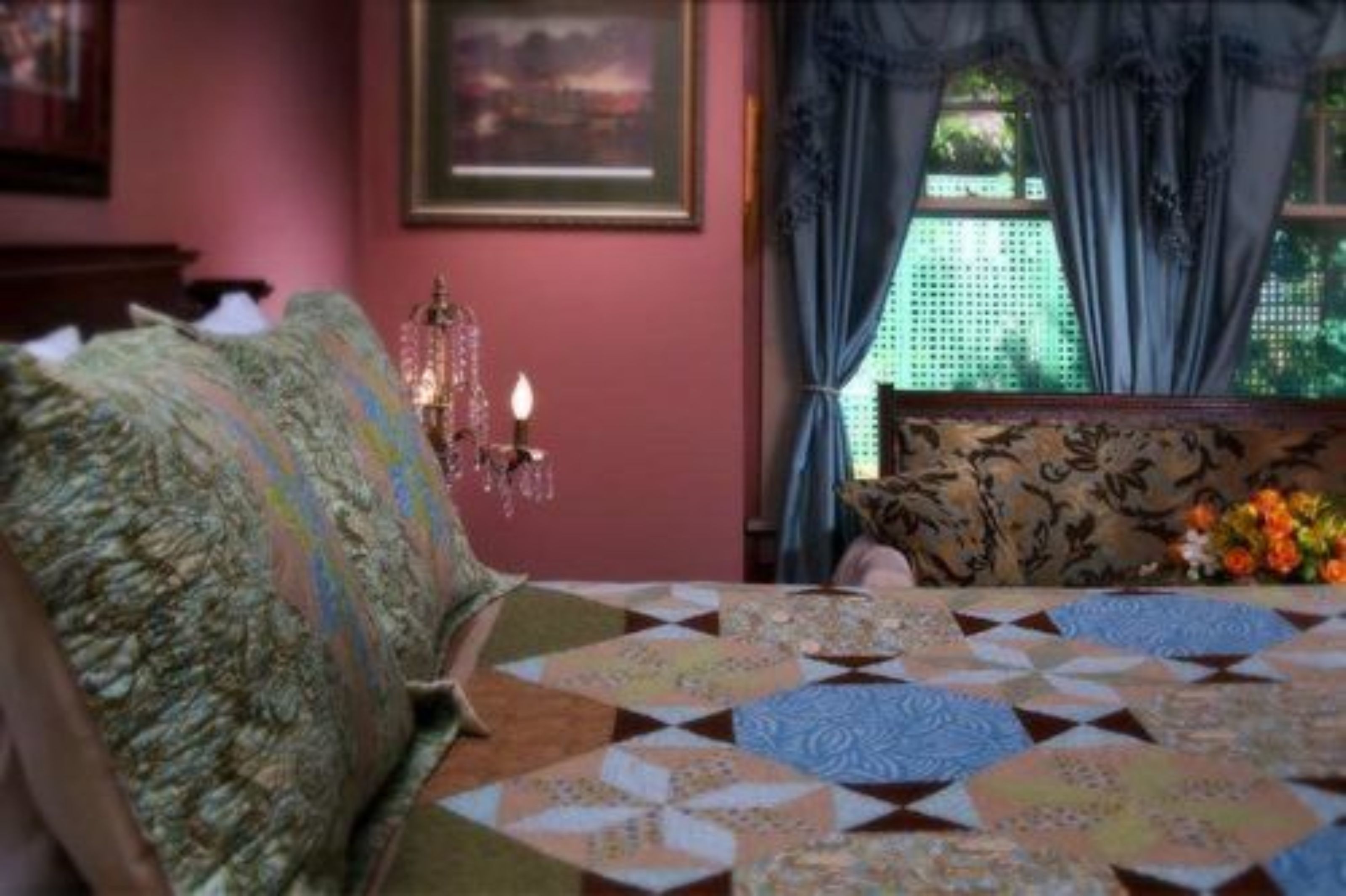 A bedroom with a patterned bedspread, a chandelier, and a view of greenery through the window.
