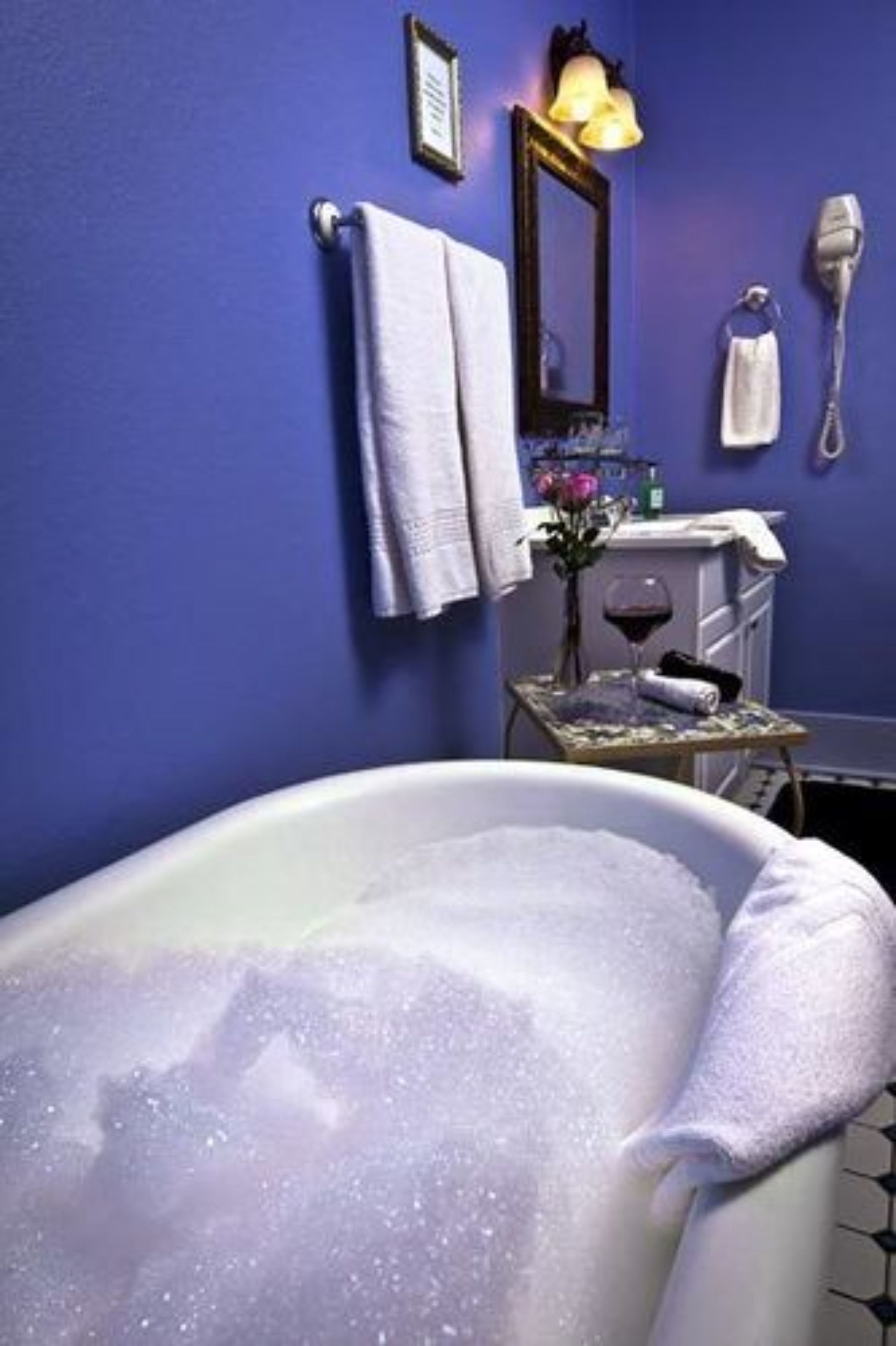 Un baño con bañera, toallero, espejo, secador de pelo y una pequeña mesa con una copa de vino y flores.