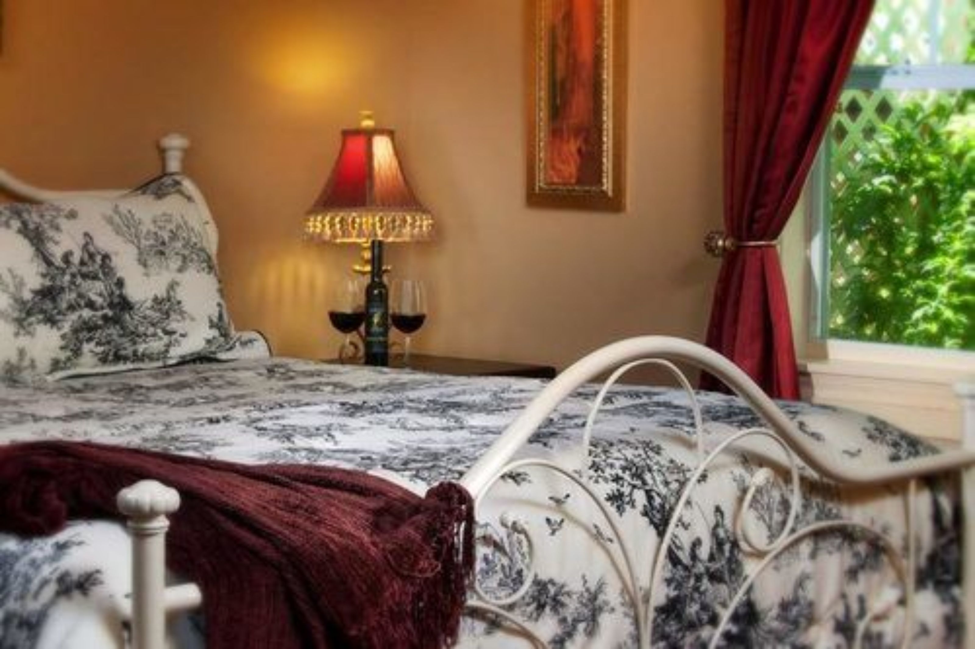 A bed with a floral bedspread, a red throw blanket, and a bedside table with a lamp and wine glasses.