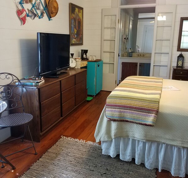 Quarto com uma cama, uma televisão sobre uma cômoda, uma cafeteira e uma porta que dá para outro espaço.