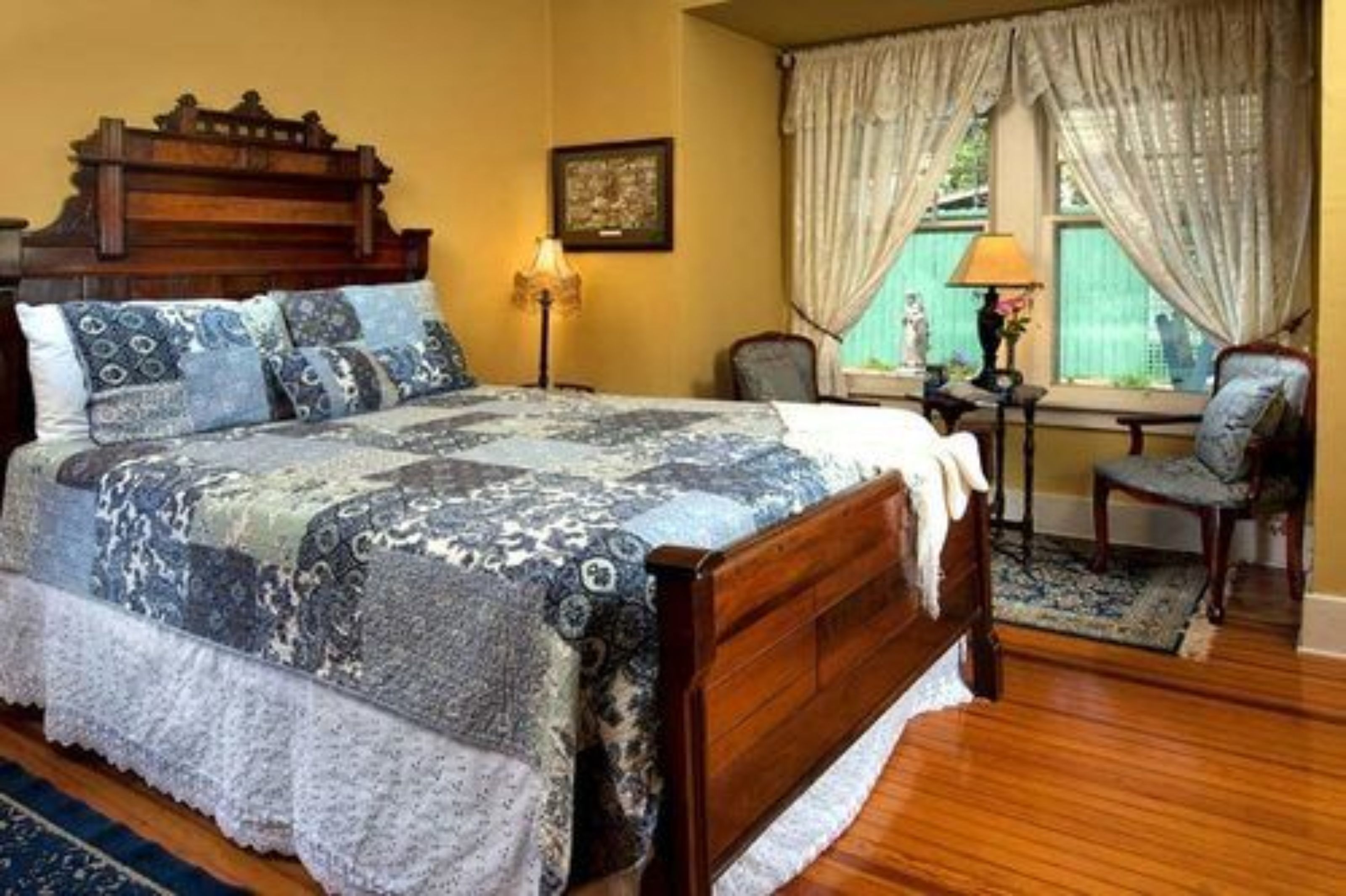 Un dormitorio con una cama de madera, ropa de cama estampada, una mesita de noche, una silla y una ventana con cortinas.