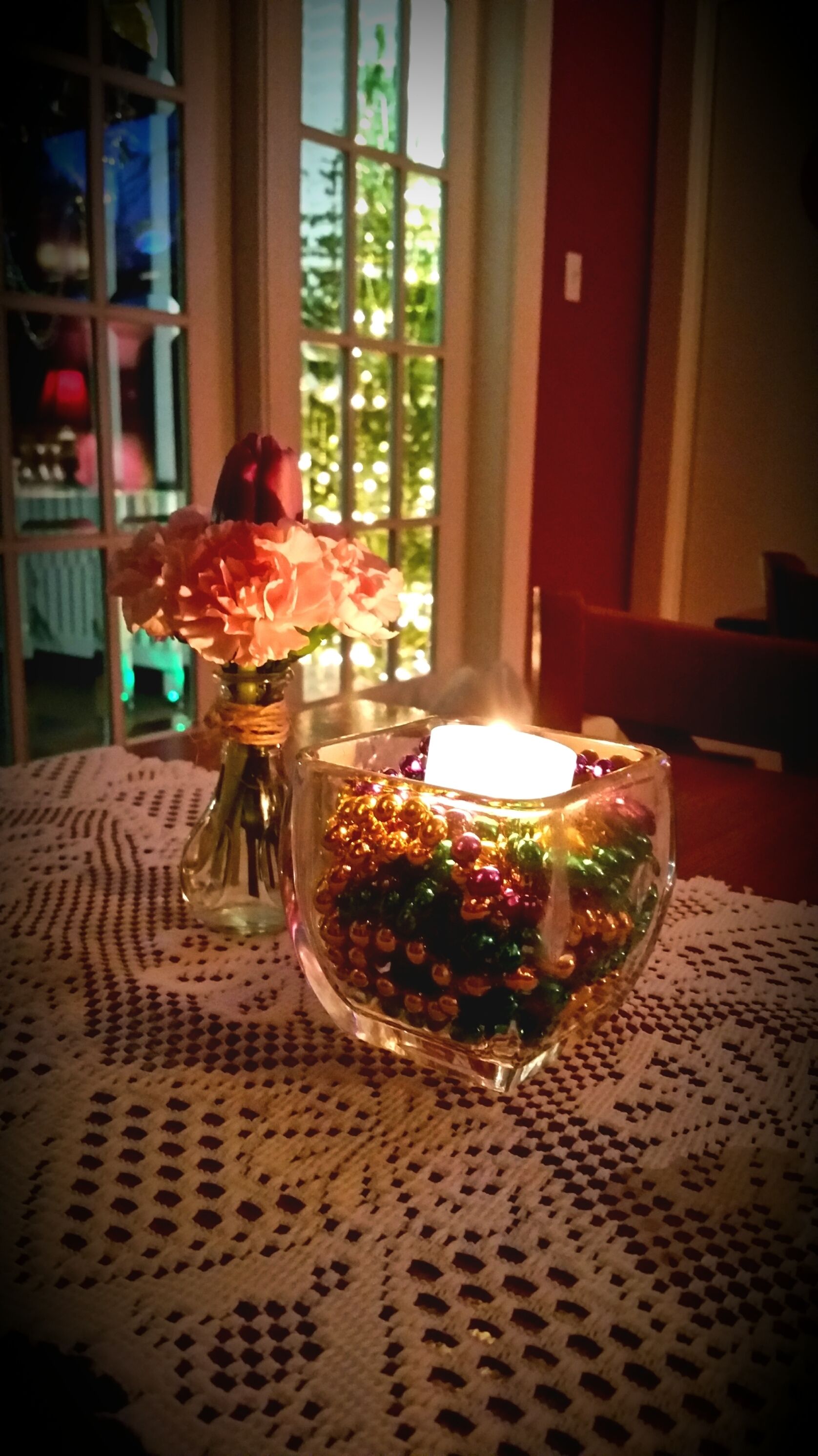 A table with a glass candle holder containing colorful beads and a vase with flowers.