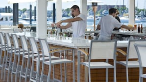 A bar area with white chairs and tables, a bartender preparing drinks, and a view of boats in the background. - Forty 1 North (Newport)