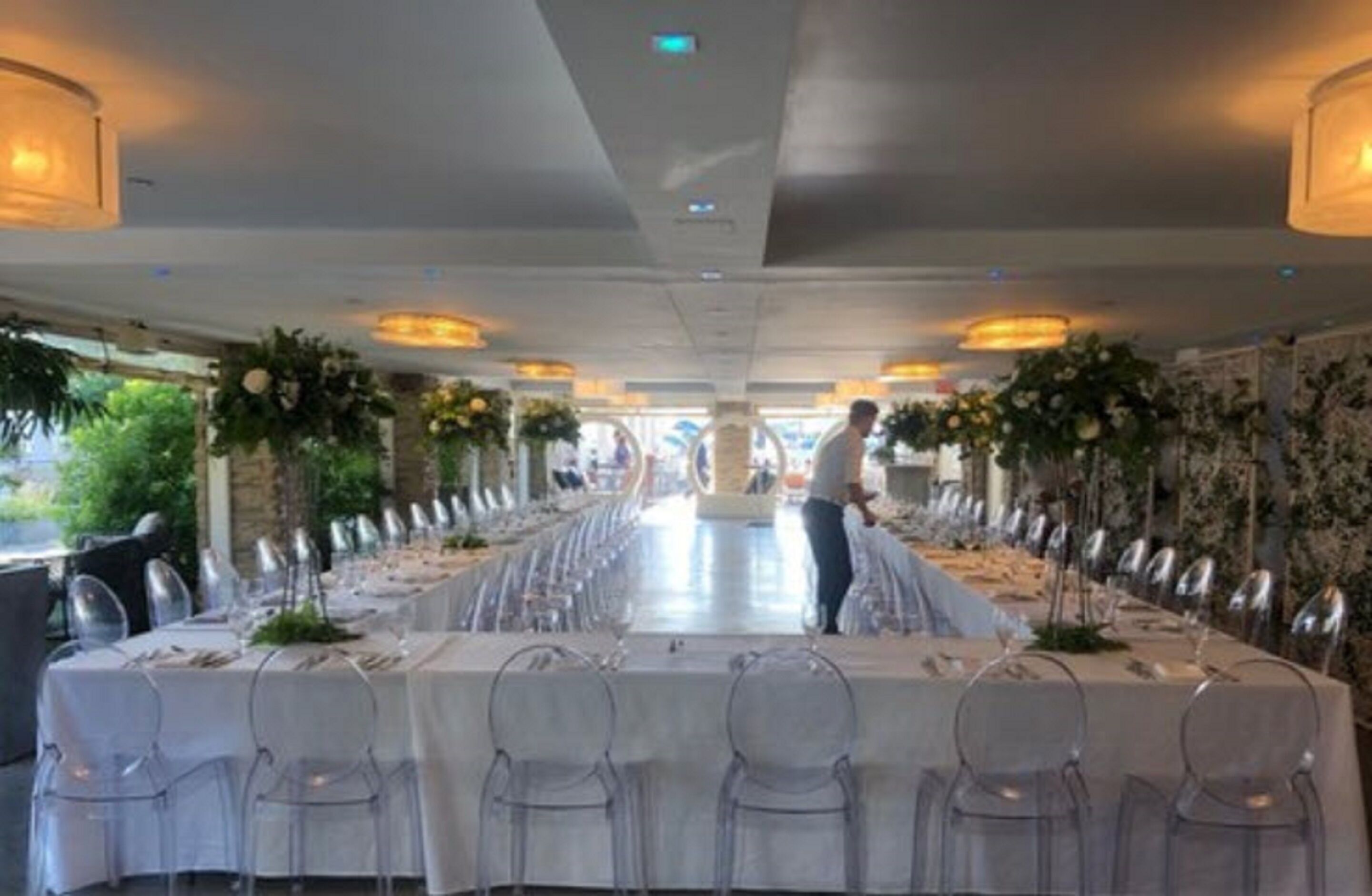 A long dining table set with chairs, centered with a person standing, and surrounded by floral arrangements.