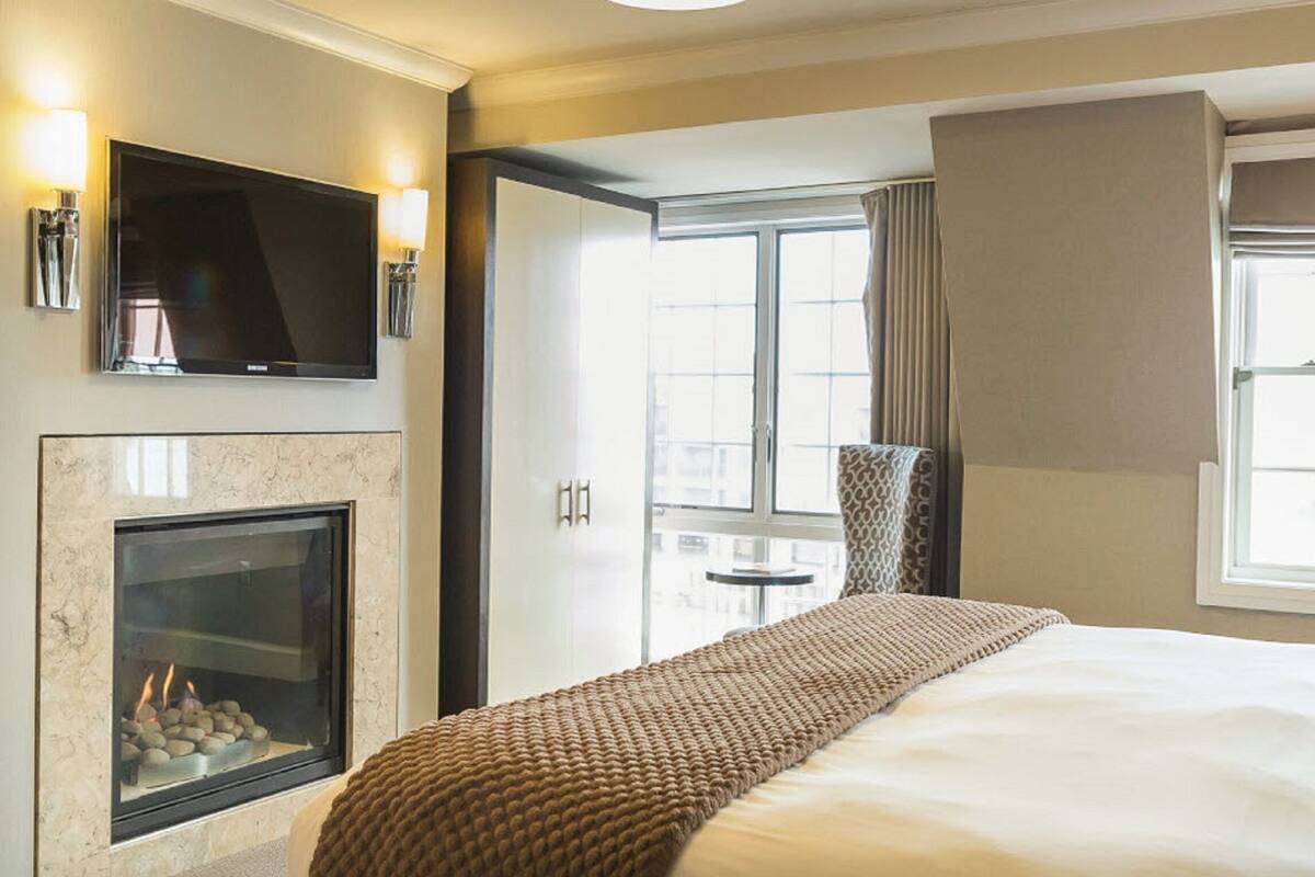 A hotel room with a large bed, a flat-screen TV mounted above a fireplace, and a window with curtains.