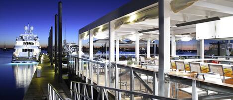 Een restaurant aan het water met terras, uitzicht op het water, met een groot schip afgemeerd in de buurt en een modern architectonisch ontwerp.