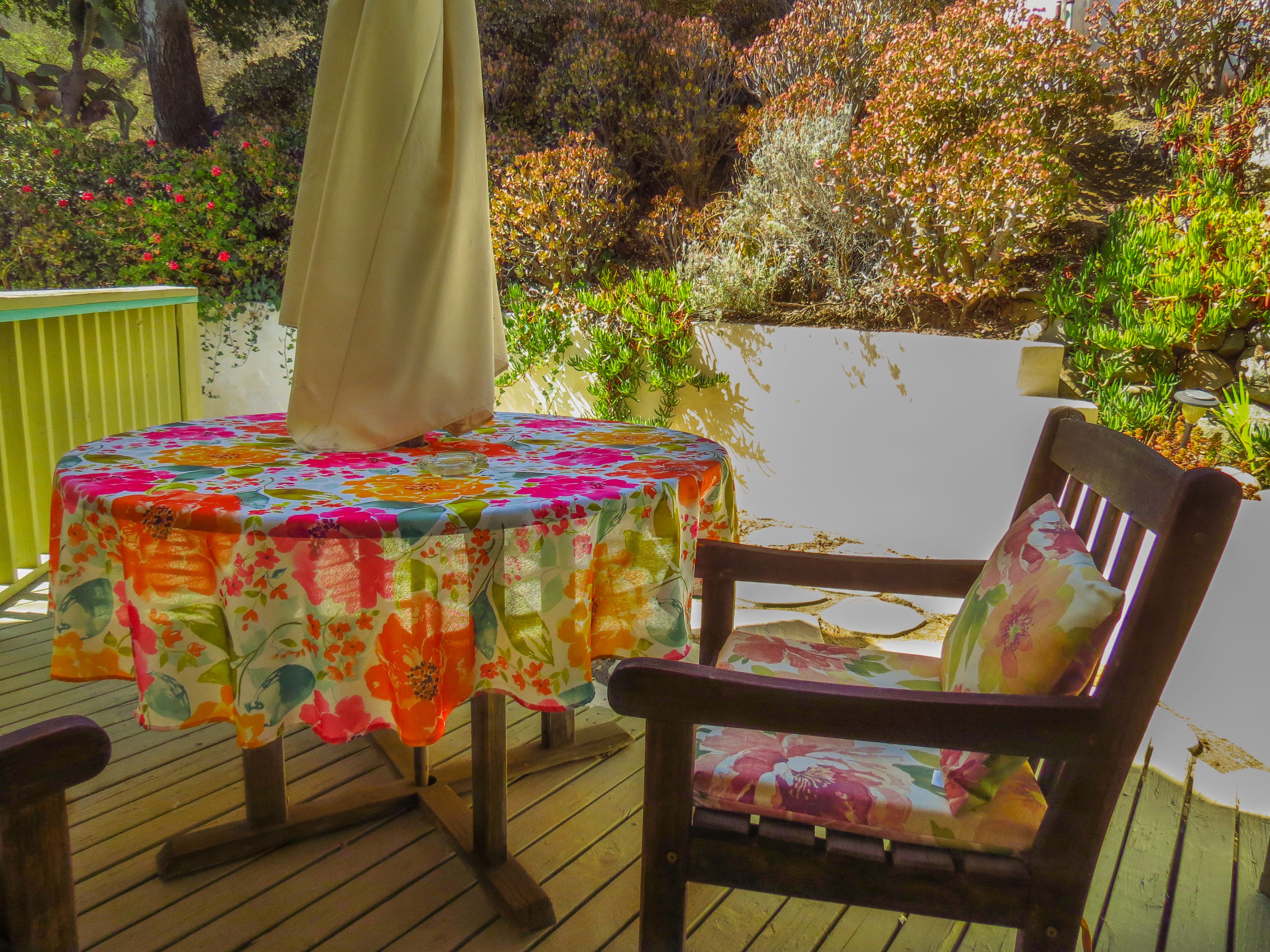 A wooden deck with a round table covered by a floral tablecloth, a wooden chair with a matching cushion, and a beige umbrella.