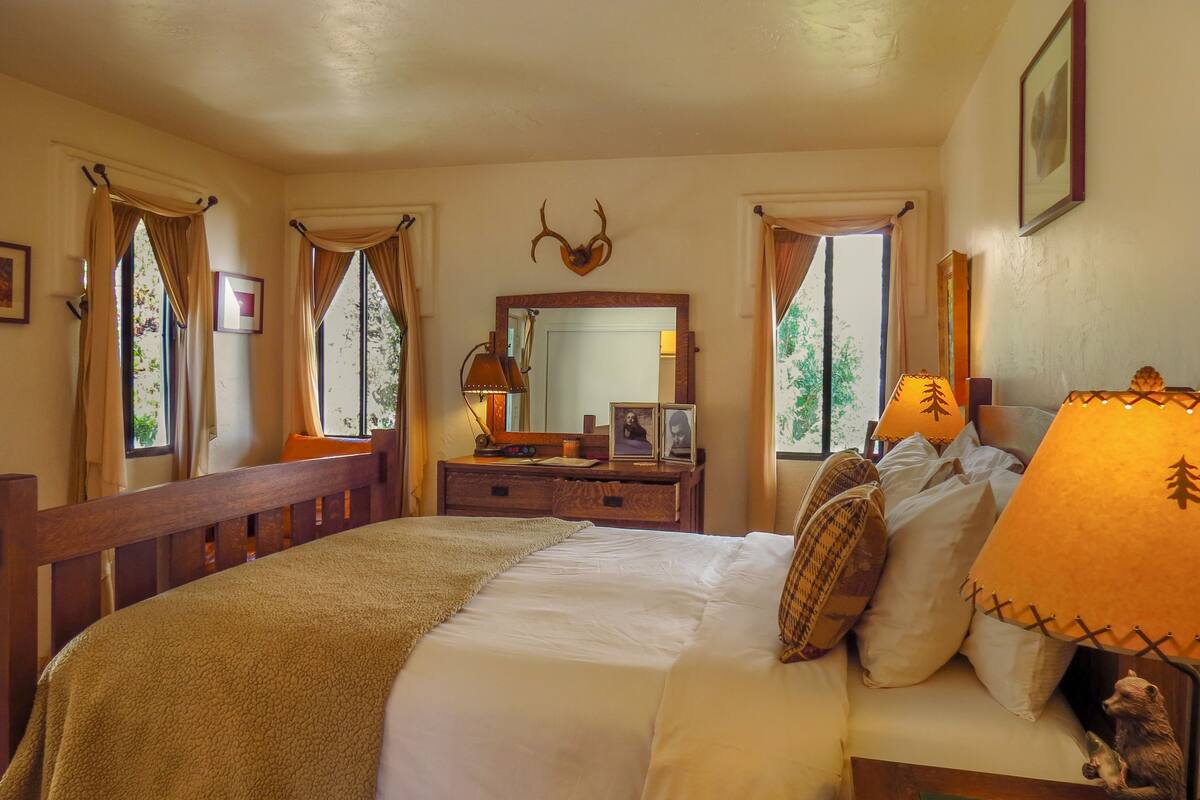 A bedroom with a large bed, a dresser, a mirror, and two windows with curtains.