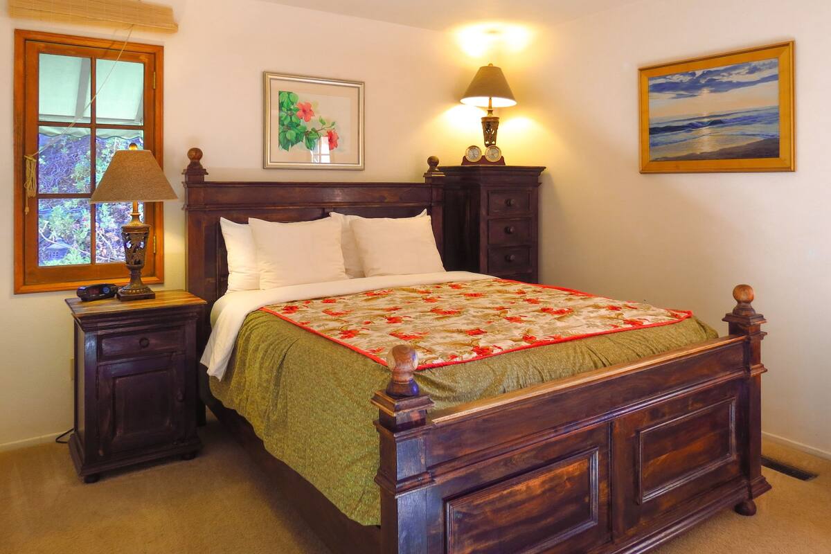 A bedroom with a wooden bed, a nightstand with a lamp, a dresser, and a window with blinds.