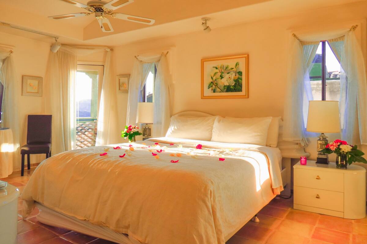 A hotel room with a large bed, a ceiling fan, a nightstand with a lamp, and a window with curtains.